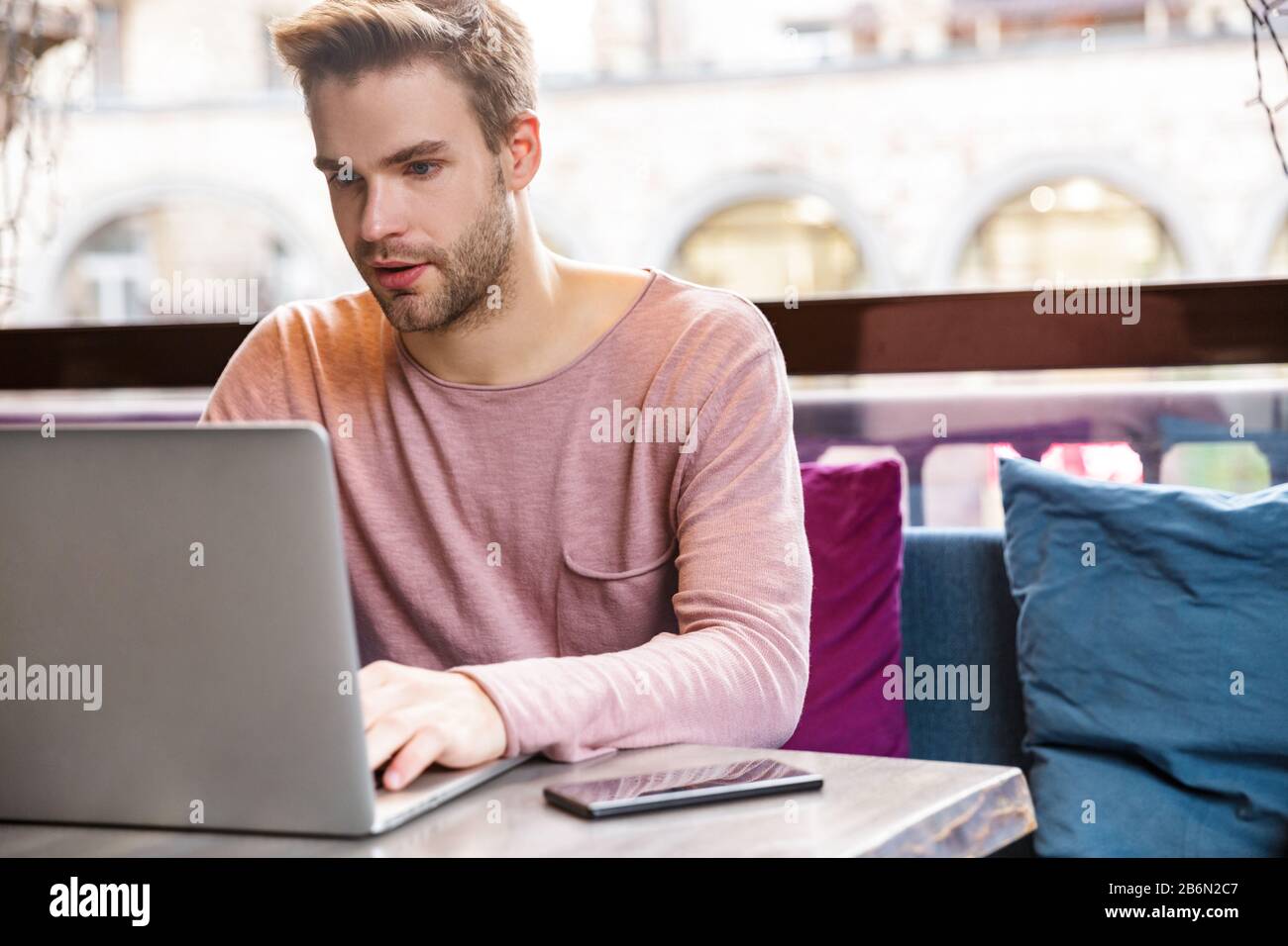 Bel giovanotto pensoso che lavora su un computer portatile mentre si siede nel caffè Foto Stock