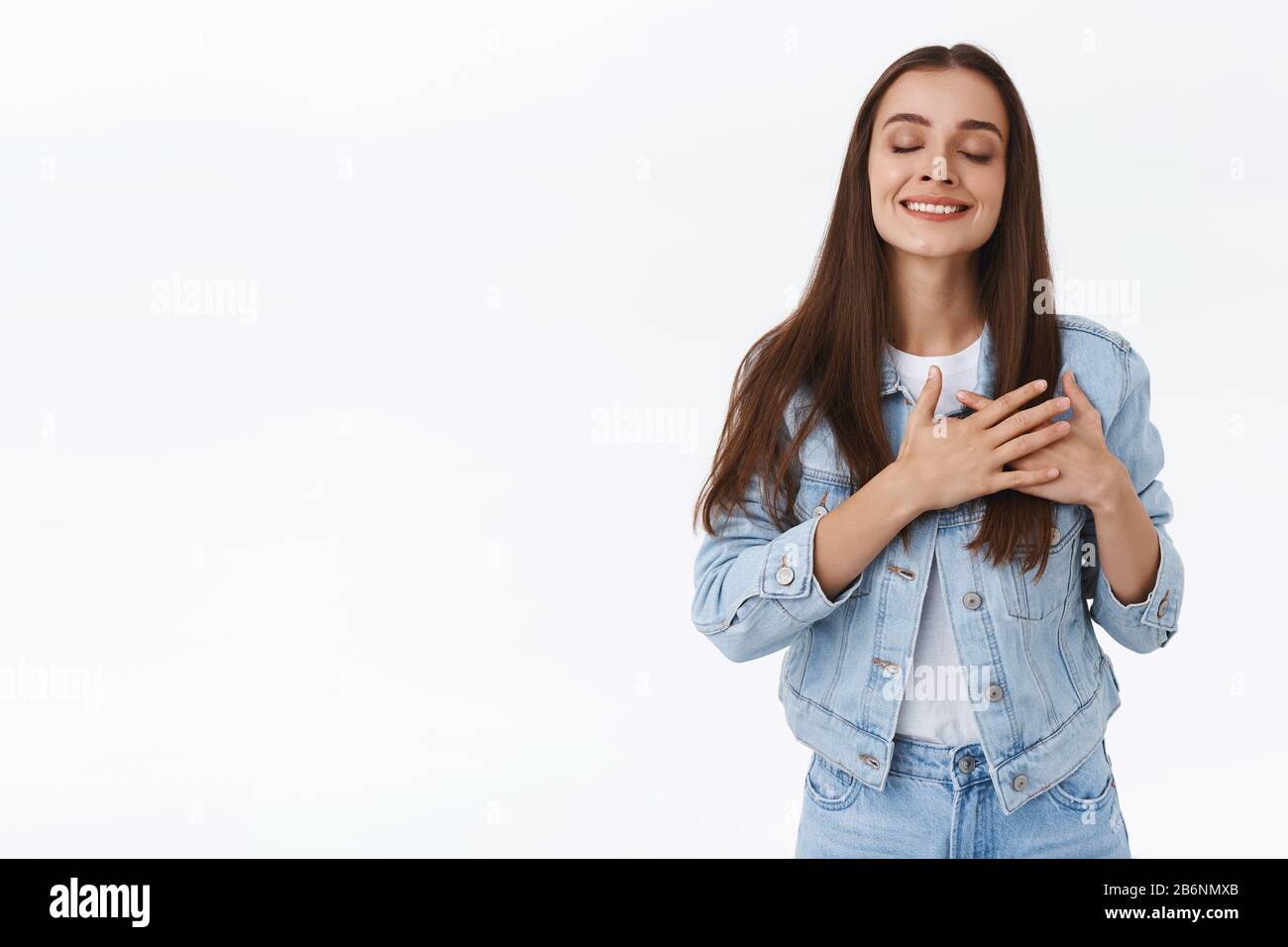 Donna caucasica in giacca di denim, jeans, che si tocca il suo cuore, gli occhi stretti e sorridendo con bella espressione, ammirare qualcuno o sentire Foto Stock