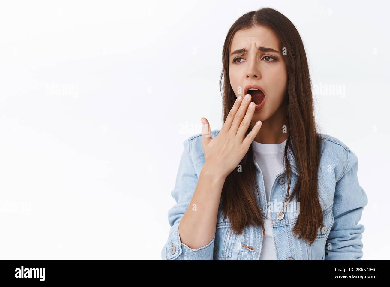 Annoiata e assonita giovane ragazza bruna stanca in giacca di denim, accigliata e sbadigia, coprire la bocca aperta, bisogno di sonno, svegliarsi presto mattina vuole Foto Stock