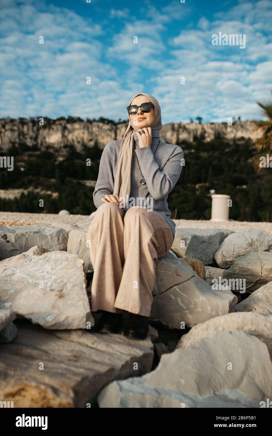 Ritratto di giovane europeo le donne musulmane con il hijab seduto sulla spiaggia di pietra con il mare sullo sfondo. Lei è felice e rilassato. Foto Stock