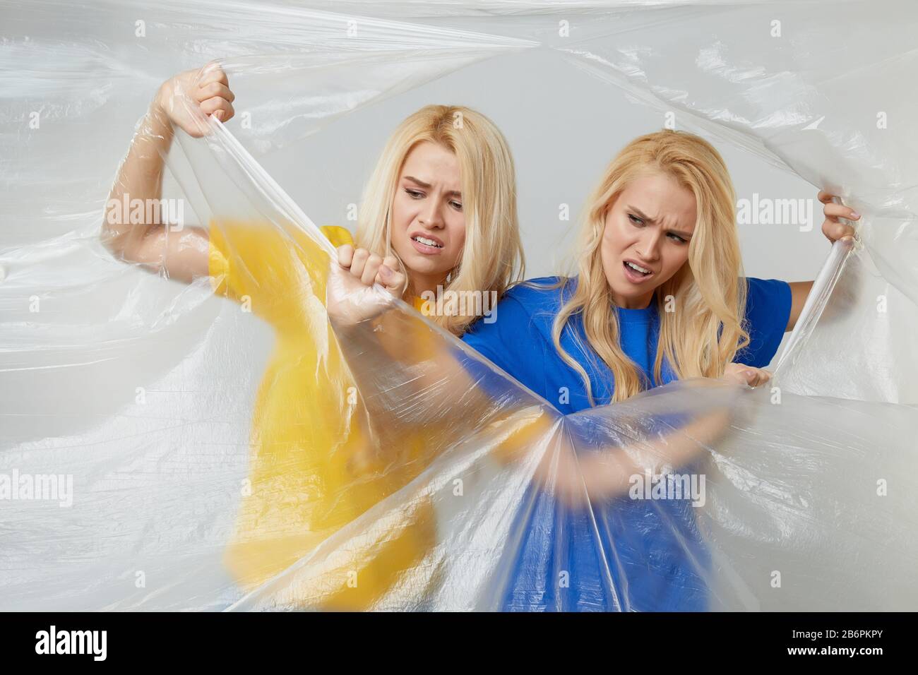 Giovani donne volontarie in giallo e blu t-shirt behing un muro di non-degradable polyethylene wrap cercando di combattere cellophane. Conce. Riciclaggio rifiuti Foto Stock