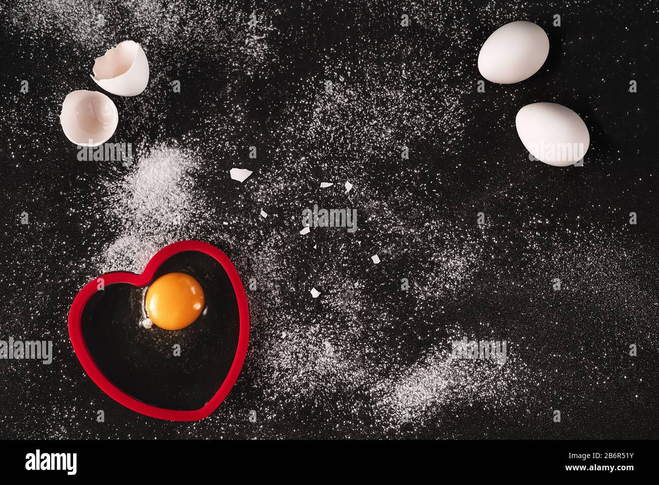 Immagine chiave bassa di un bianco crudo uova biologiche su sfondo nero cosparso di farina. Un uovo rotto aperto sulla forma rossa a forma di cuore di silicone. Cucina Foto Stock