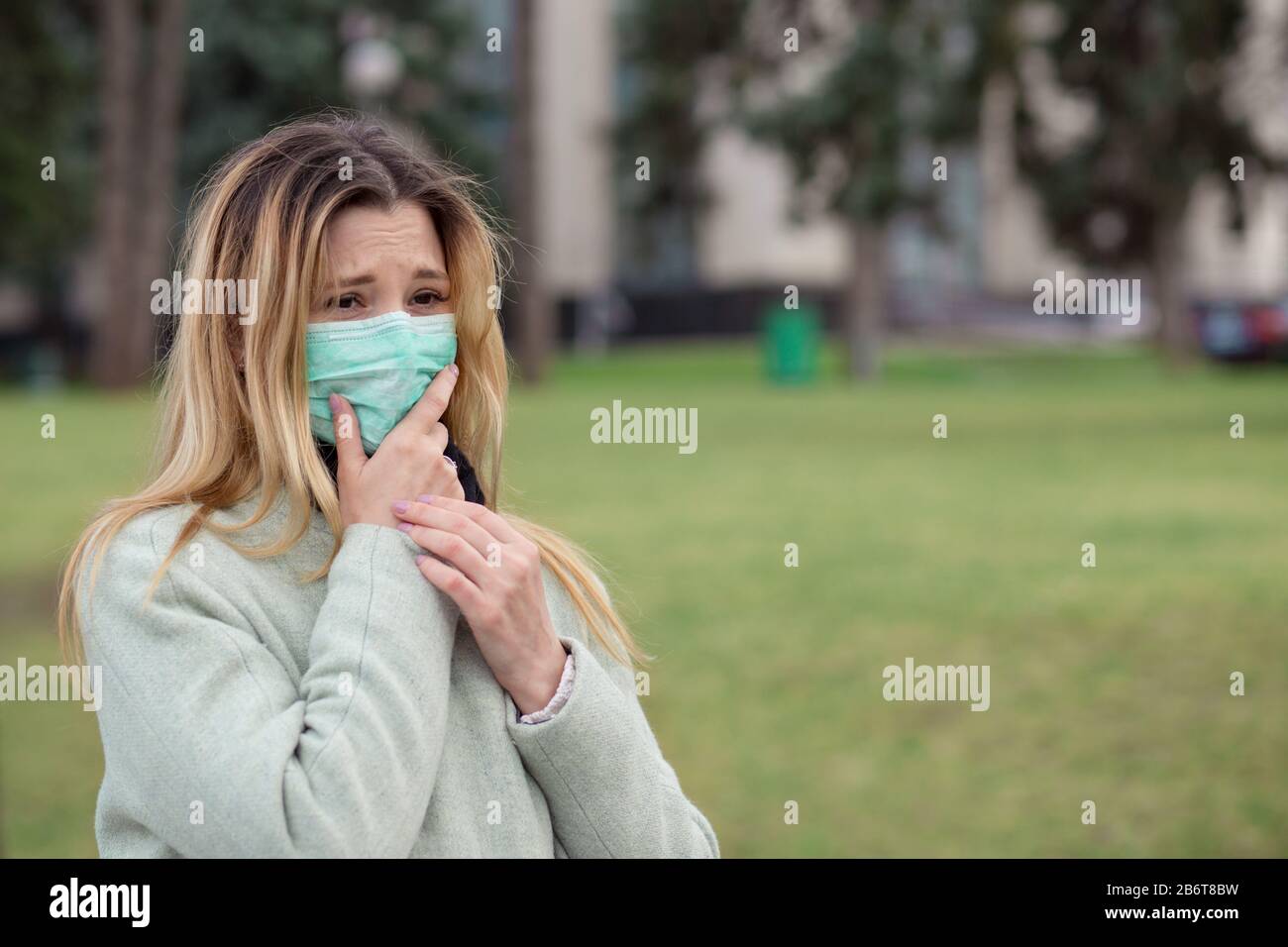 Ragazza ammalata, una giovane donna bionda in profilo che indossa chirurgia maschera medica sensazione di male, male, essere colpito dal coronavirus, Covid2019 in piedi all'aperto in Foto Stock