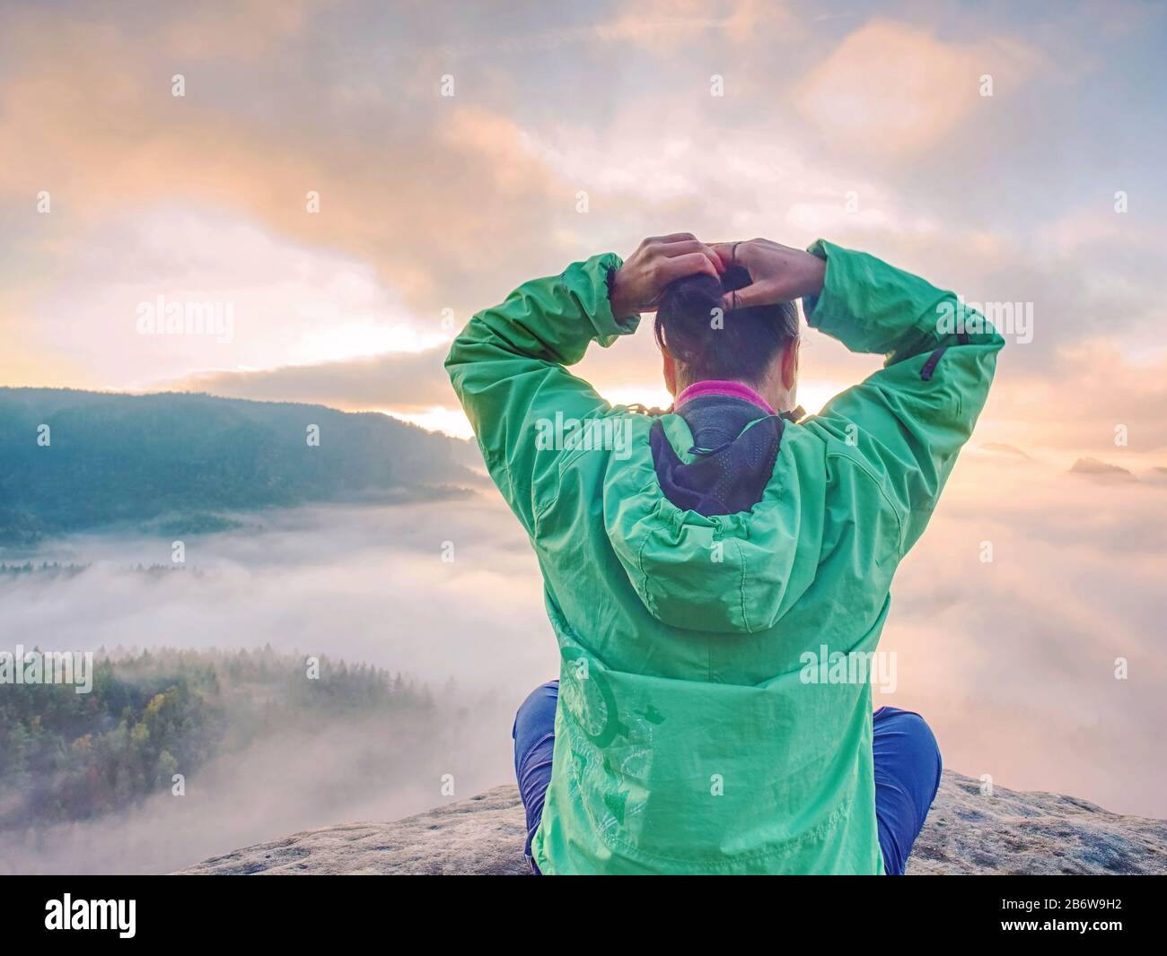 Giovane donna sedersi sulla cima di una montagna celebrare la giornata. Signora giovane escursionista in seduta verde blu giacca a vento con red zaino sulla scogliera taglienti Foto Stock