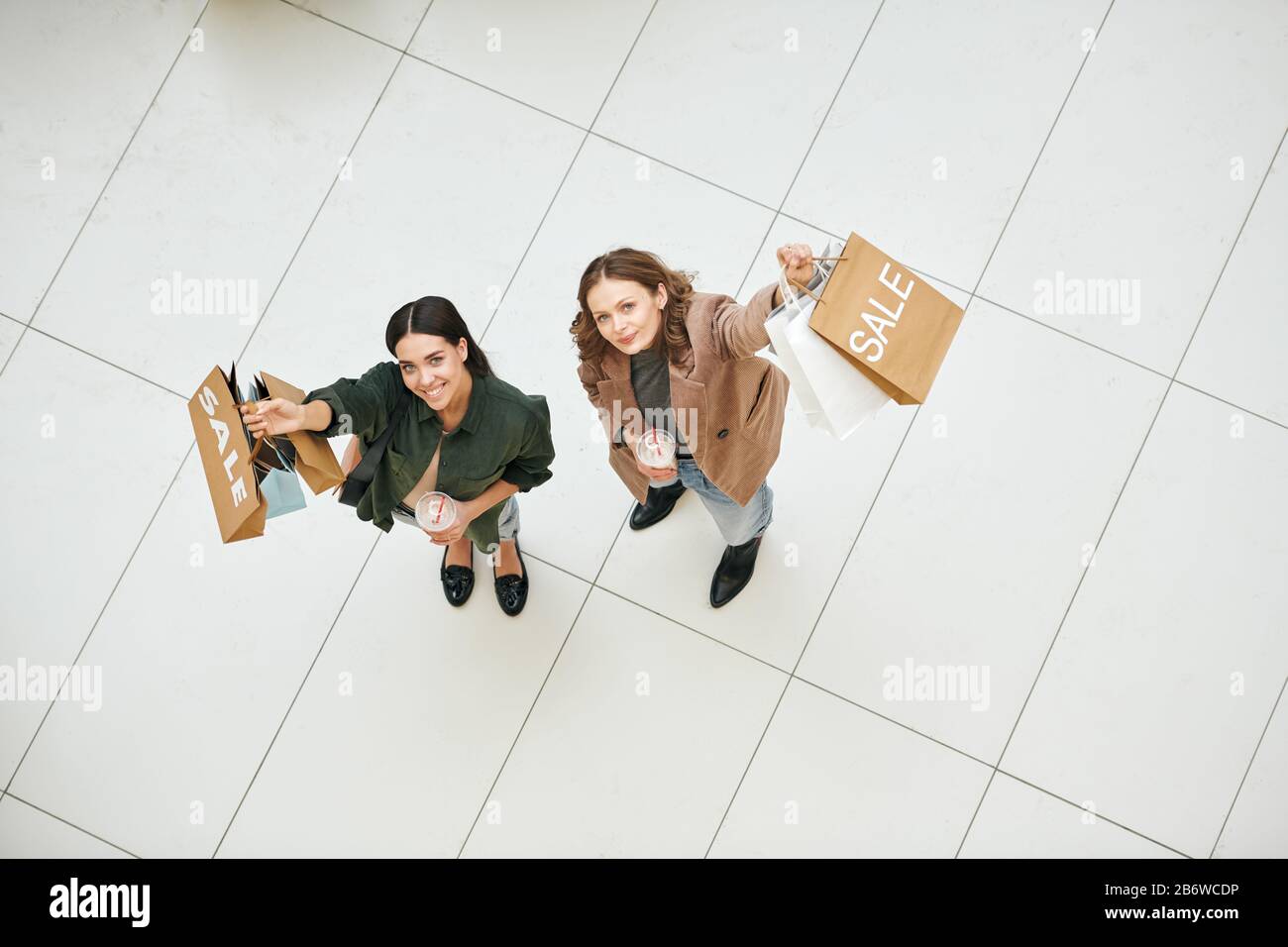 Sopra la vista delle donne positive che ondolano le borse di shopping e che guardano la macchina fotografica nella lobby del centro commerciale Foto Stock