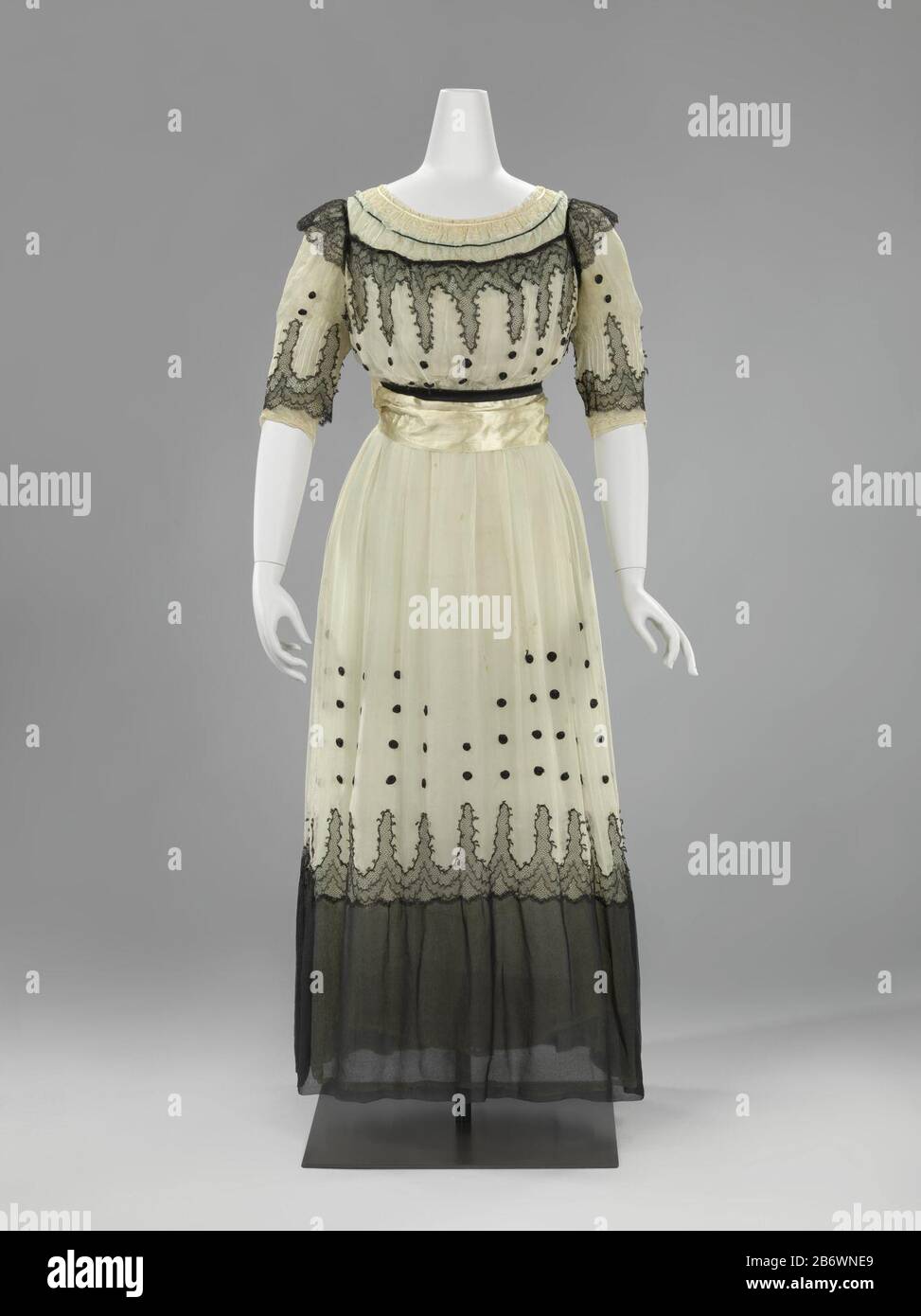 De japon is opgebouwd uit drie over elkaar liggende lagen crêpe georgette; een witte onderjurk, een 'eau de nil' japon en een wit met zwarte japon. Hierdoor heedft het geheel een blauwgroene gloed gekregen. Opvallend eigentijds zijn de cirkels van opgerold zwart satijnband op de witte rok en de zwart-witte geknopte franje aan de ceintuur. De rok è voetvrij en ruim genoeg voor voldoende bewegingsvrijheid. Produttore : anoniemoniemPlaats fabbricante: Nederland (mogelijk)Frankrijk (mogelijk) datazione: CA. 1916 - ca. 1919 kenmerken Fisico: crêpe georgette, machinale kant, machinale Chantilly Foto Stock