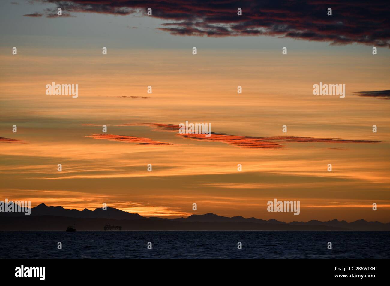 Stagionatura. Cielo rosso all'alba, piattaforma di perforazione a olio iт Baia di Mossel, silhouette di montagne all'orizzonte. Sudafrica. Foto Stock