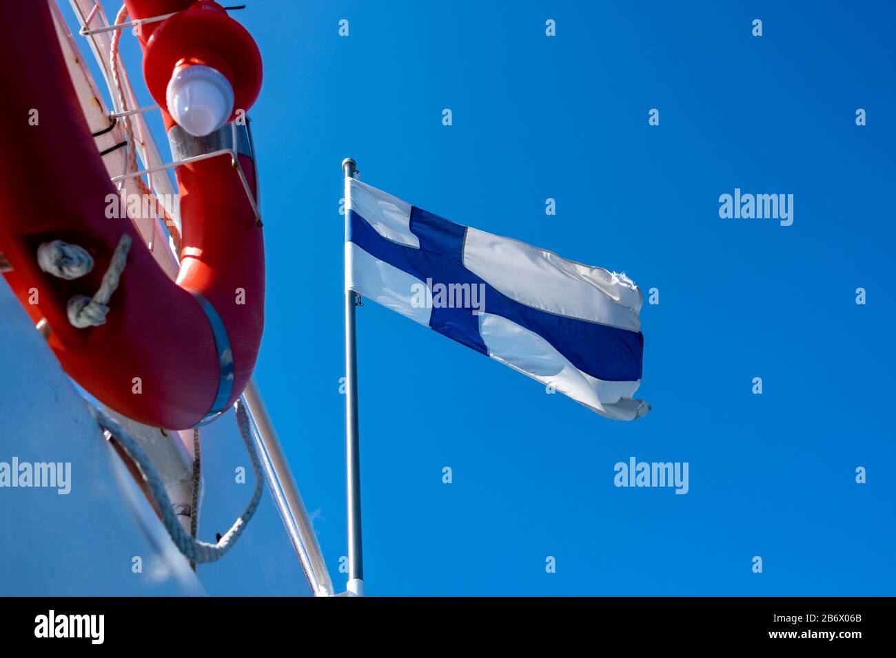 Bandiera finlandese che sventola nel vento contro il cielo blu. La lifebuoy arancione è in primo piano. Foto Stock