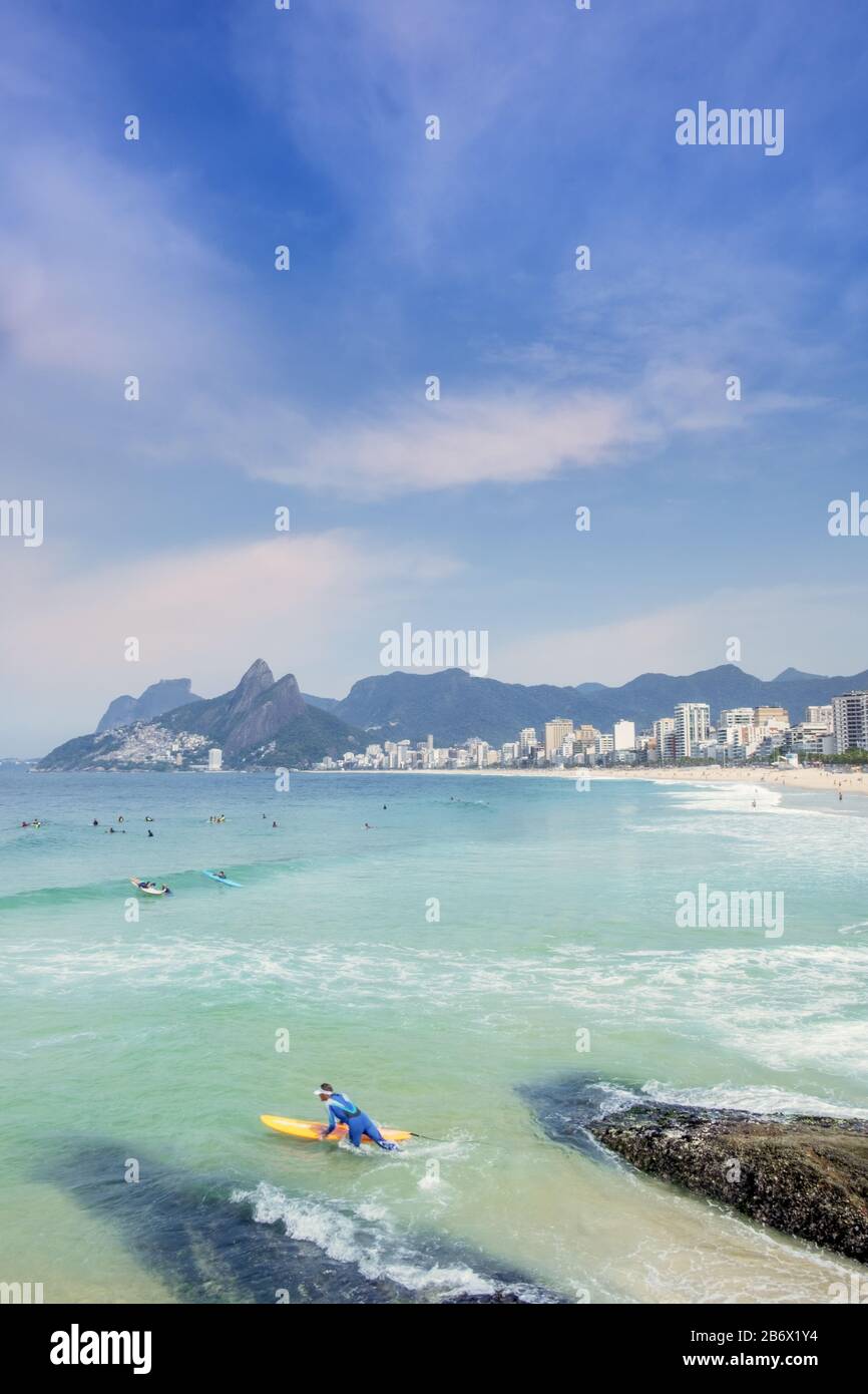 Un surfista maschio che entra nell'Oceano Atlantico alle rocce Arpoador con spiaggia Ipanema, quartiere e le montagne Dois Irmaos sullo sfondo Foto Stock