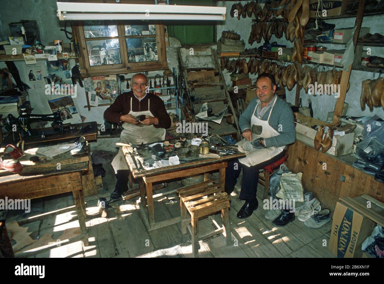 Affollato interno di un vecchio negozio di ciottoli a Metsovo, Epiro, Grecia. I due produttori di scarpe lavorano con scarpe e attrezzi appesi intorno a loro. Foto Stock