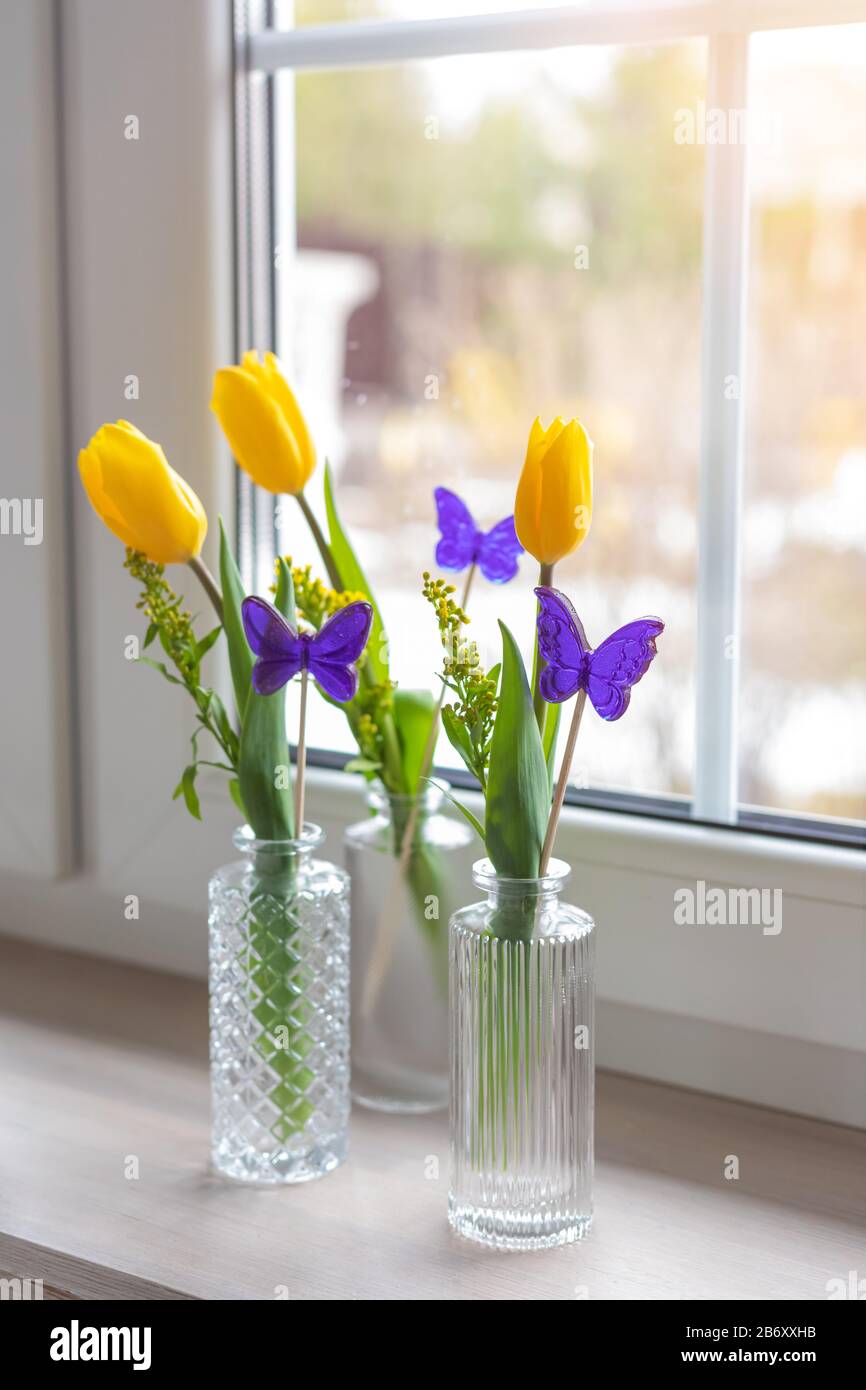Composizione primaverile con tulipani freschi gialli e caramelle a forma di farfalla viola in vasi di vetro sul davanzale, 8 marzo, Pasqua, Madre e Wome Foto Stock