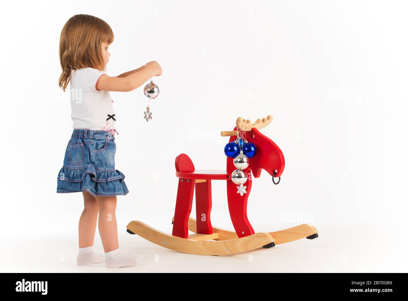 Piccola ragazza affascinante gioca con un alce di legno una sedia a dondolo e decora i giocattoli di Capodanno su uno sfondo bianco in studio. Concetto di Capodanno Foto Stock