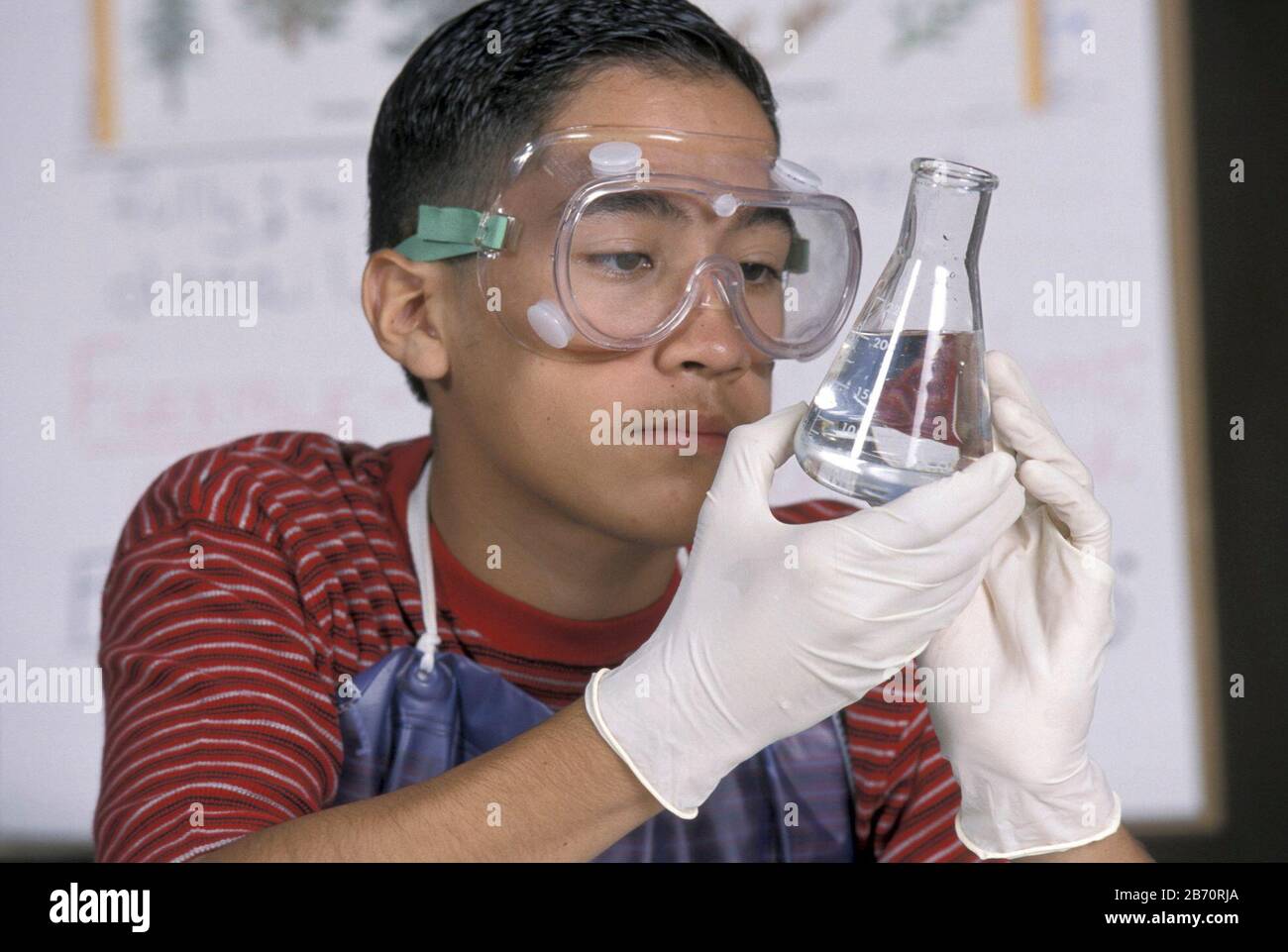 Lo studente ispanico maschio nella classe di scienza della scuola media indossa occhiali di sicurezza e guanti protettivi durante l'esperimento. ©Bob Daemmrich Foto Stock