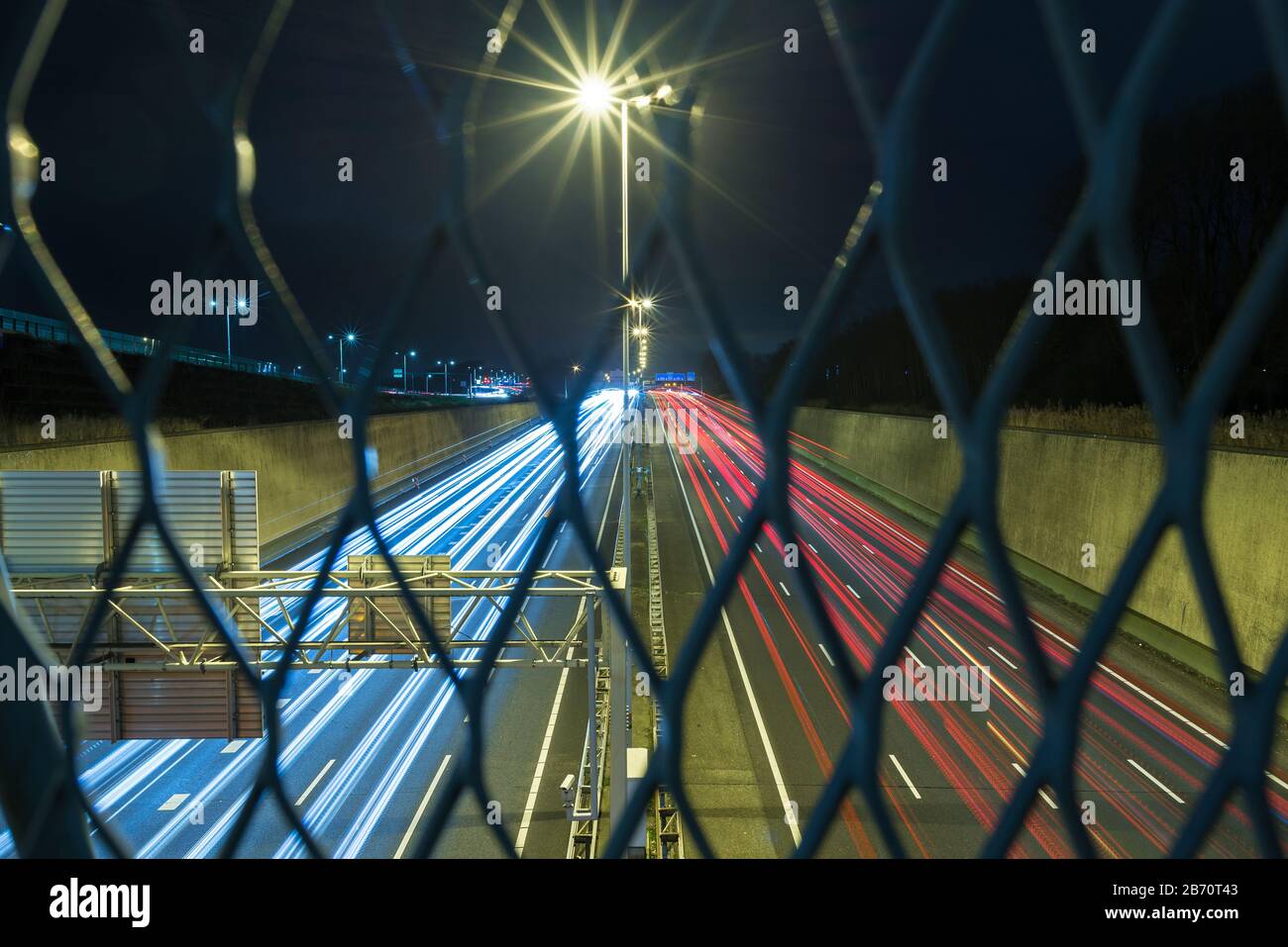 Piste per auto leggere su un'autostrada nei Paesi Bassi di notte. Vista da dietro una recinzione di metallo. Messa a fuoco selettiva. Foto Stock