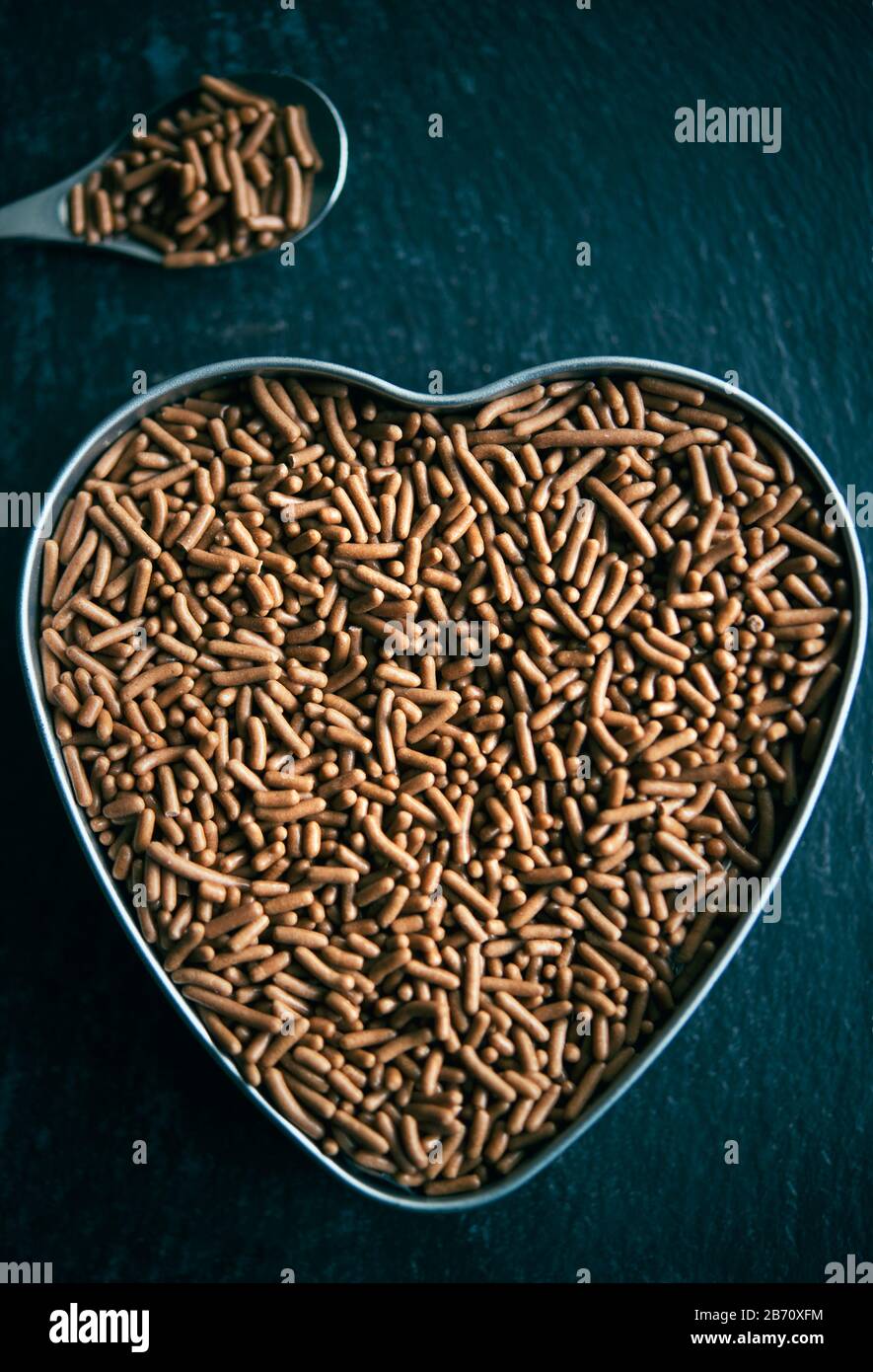 Il cioccolato viene spruzzato in una piccola teglia a forma di cuore accanto ad un cucchiaio d'argento, anch'esso pieno di pezzi di cioccolato Foto Stock