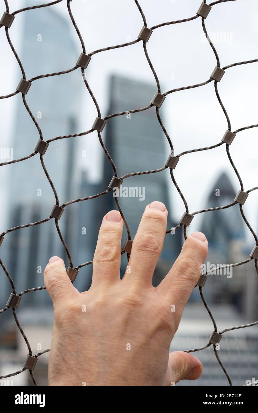Primo piano della mano dell'uomo sulla rete metallica che cerca di raggiungere gli edifici della City of London sullo sfondo come concetto di ansia e incertezza urbana Foto Stock