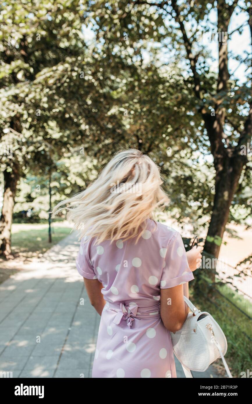 Bella donna bionda in abito longuette punteggiato polka nel parco estivo della città. È felice. Indossa una piccola borsa bianca e occhiali da sole neri. Foto Stock