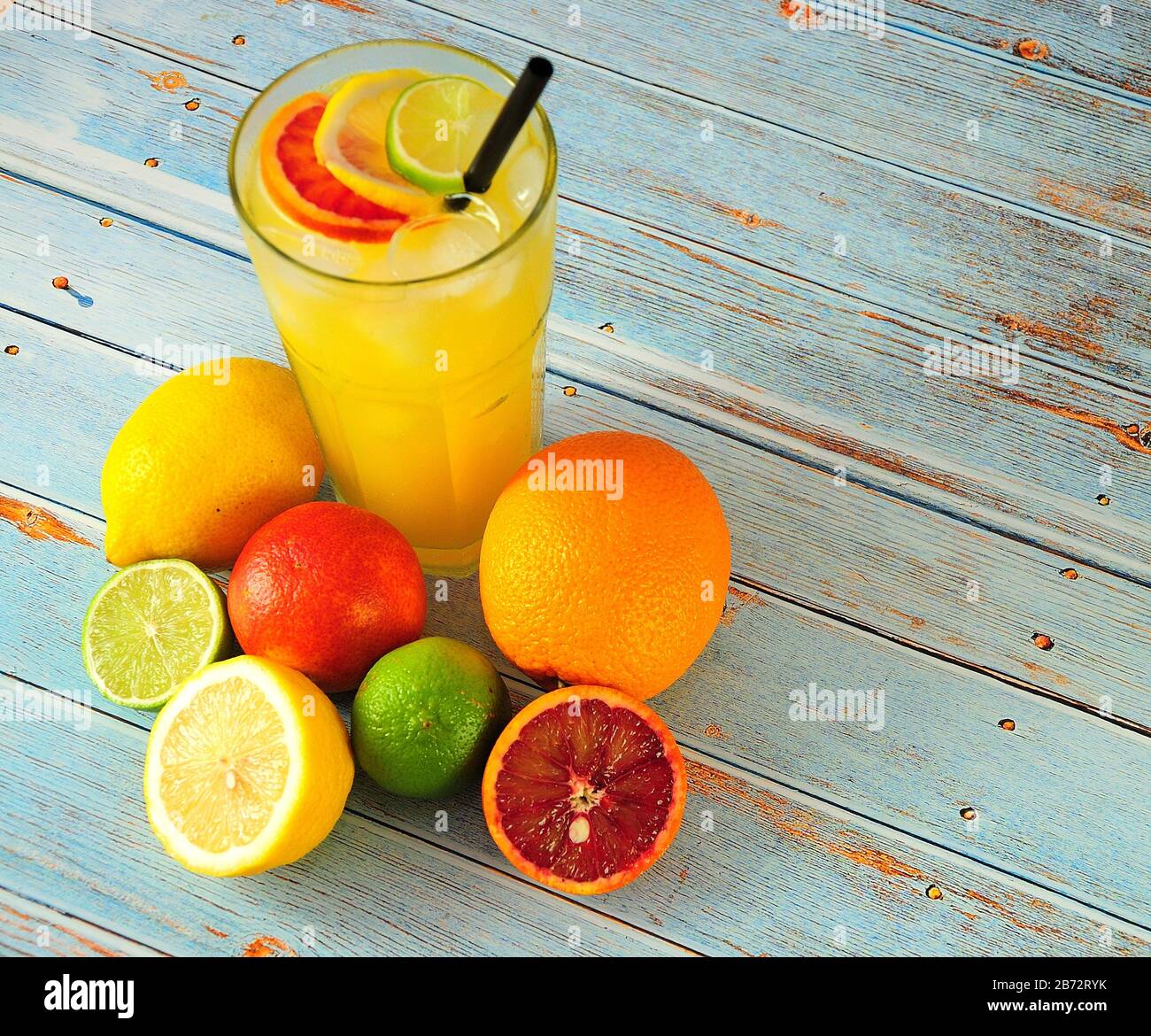 Un'alta tazza di vetro di succo di agrumi con ghiaccio e paglia, si erge su un tavolo in un mucchio di arance, limoni, lime e un piccolo pompelmo interi e tritati. Foto Stock