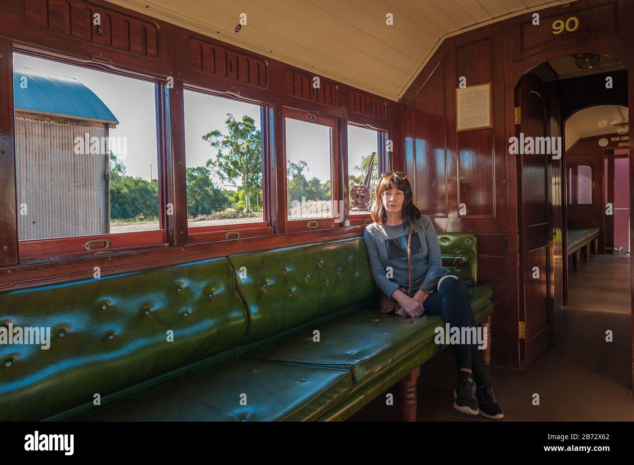 Lone femmina turista seduto su un sedile panchina verde imbottito su una vecchia carrozza di legno in attesa della partenza dei treni dalla stazione di Quorn in Australia Meridionale. Foto Stock