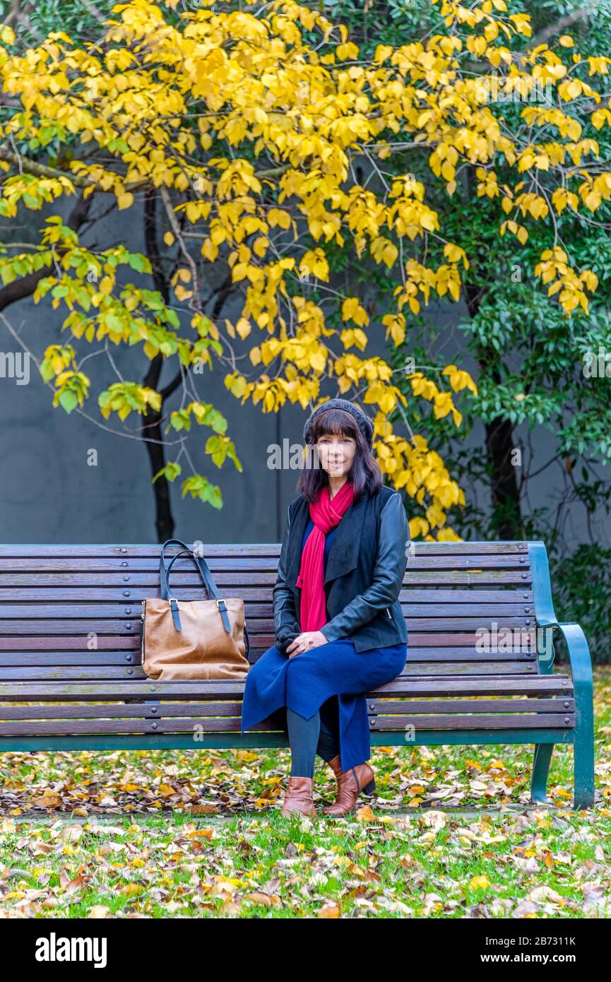 Vista del turista femminile con sciarpa rossa e borsa in pelle seduta su una panchina del parco sotto gli alberi autunnali a Fitzroy Park, Melbourne, Victoria. Foto Stock