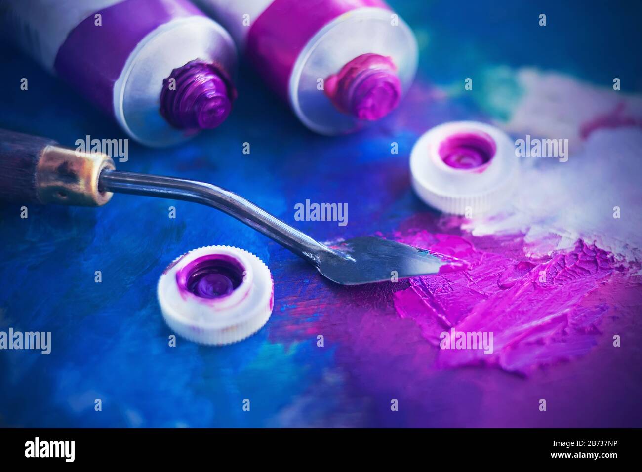 Due tubi aperti di vernice ad olio rosa e viola giacciono accanto ad un coltello per palette su una tavolozza dipinta in diversi colori. Foto Stock