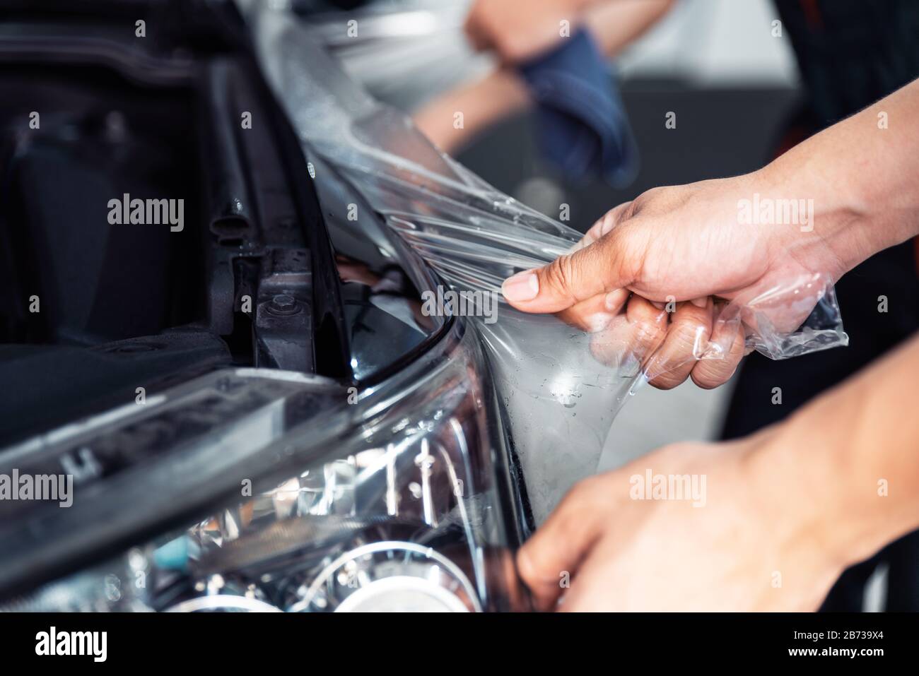Processo di installazione PPF, stretching ravvicinata, rimozione manuale di una vecchia pellicola trasparente su un faro e cofano dell'auto. Servizio professionale automobilistico. Foto Stock