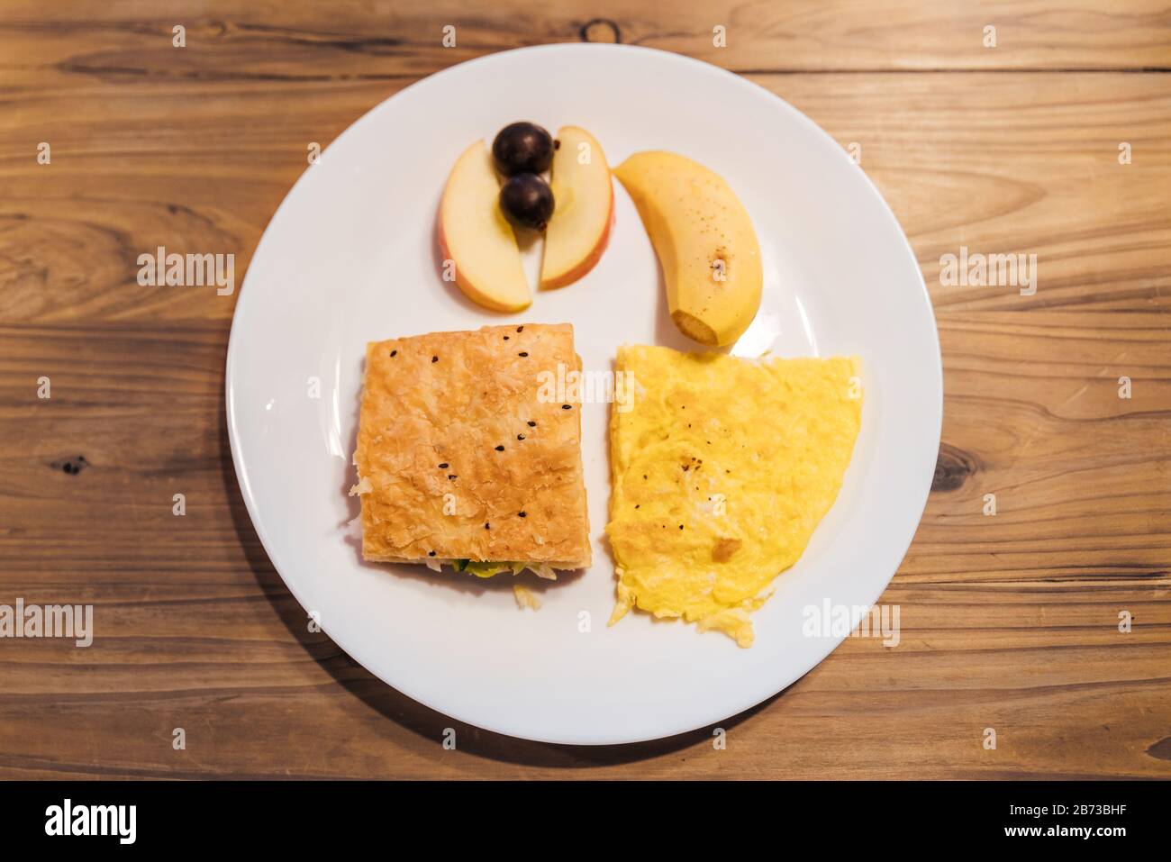 Vista dall'alto del menu colazione: Omelette, sandwich croissant, banana, mela e uva in piatto bianco su tavolo di legno. Taipei ostello servizio alimentare. Foto Stock