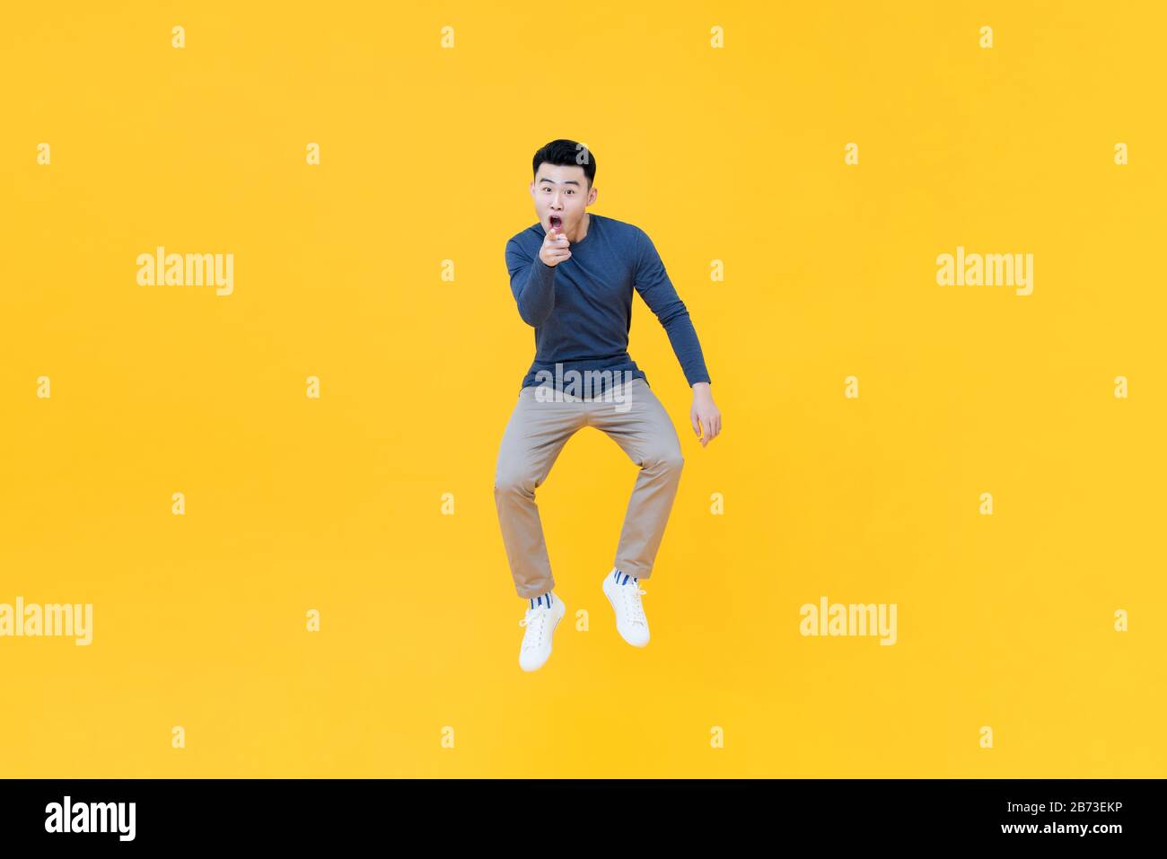 Ritratto a tutta lunghezza di giovane felice uomo asiatico che salta a mezz'aria mentre in un divertente gesto di puntamento su sfondo giallo isolato studio Foto Stock