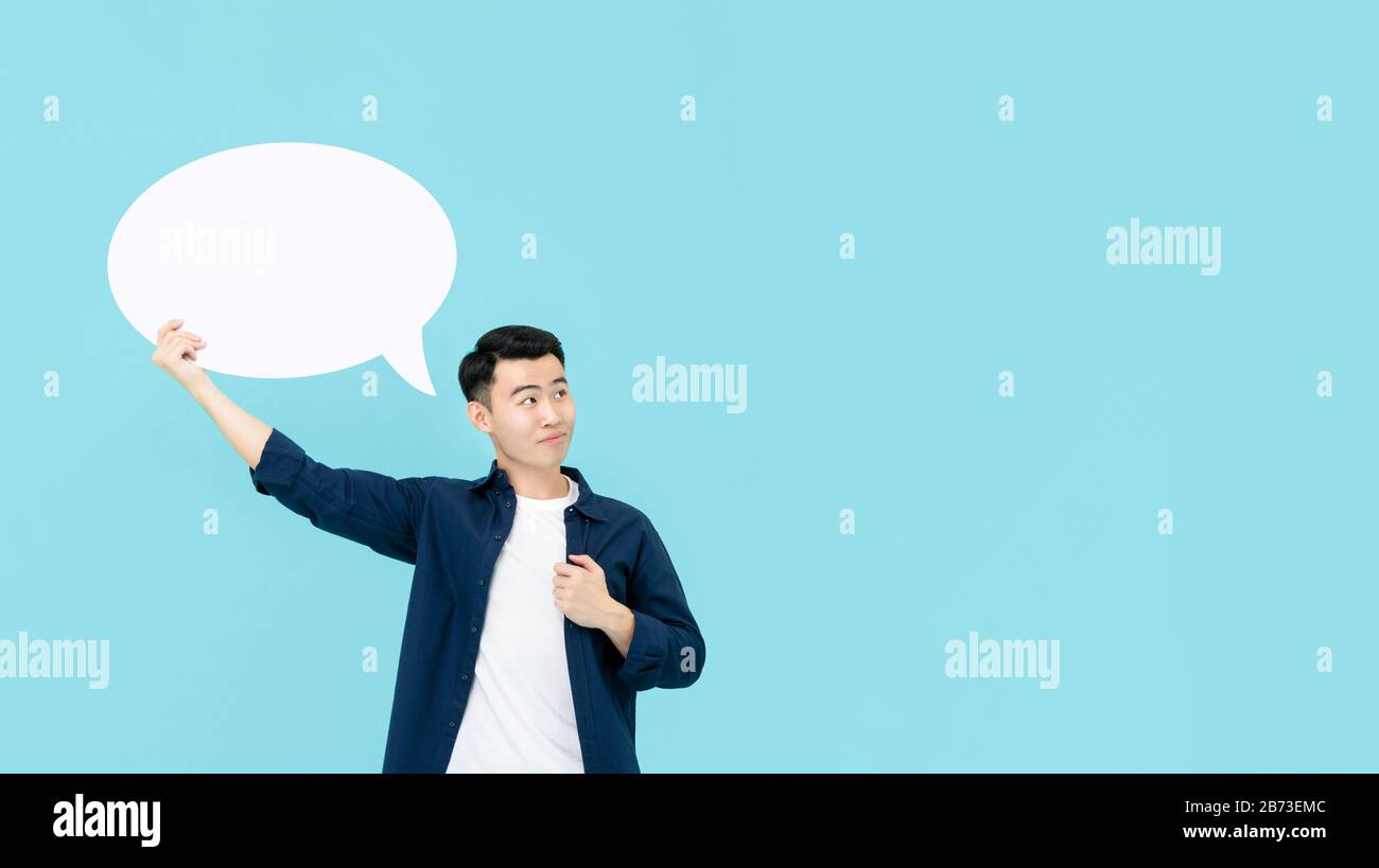 Giovane uomo asiatico sorridente che tiene vuoto bianco discorso bolla whlie pensare e guardare lo spazio di copia in chiaro blu studio sfondo Foto Stock