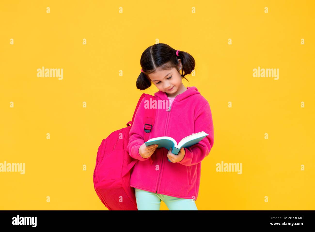 Ritratto di bella ragazza carina con capelli pigtail in giacca rosa e zaino lettura libro in giallo isolato studio sfondo Foto Stock