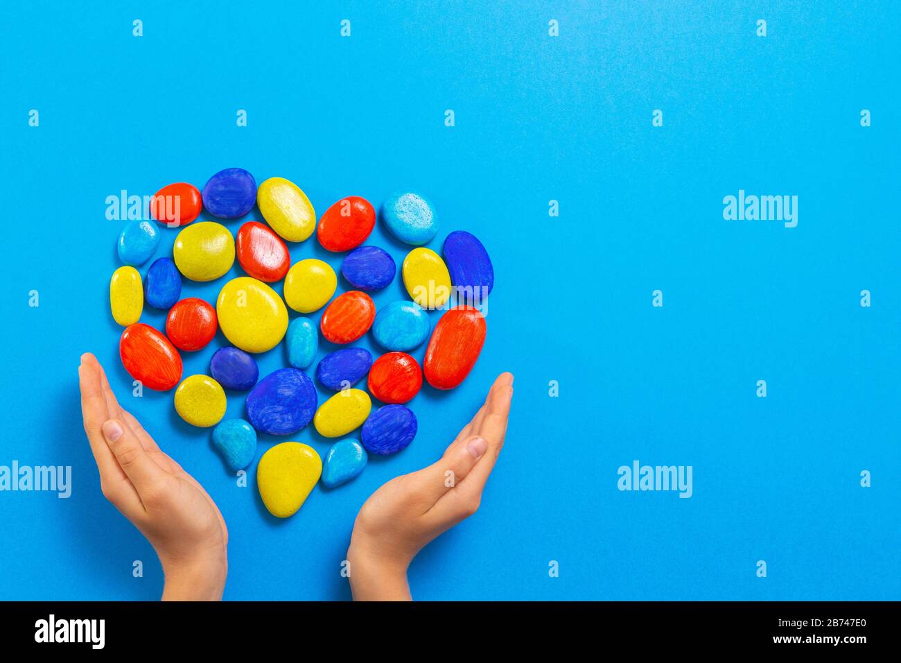 Concetto del giorno di consapevolezza di autismo del mondo. Mani del bambino con cuore fatte di pietre di colore rosso giallo blu su sfondo blu chiaro Foto Stock