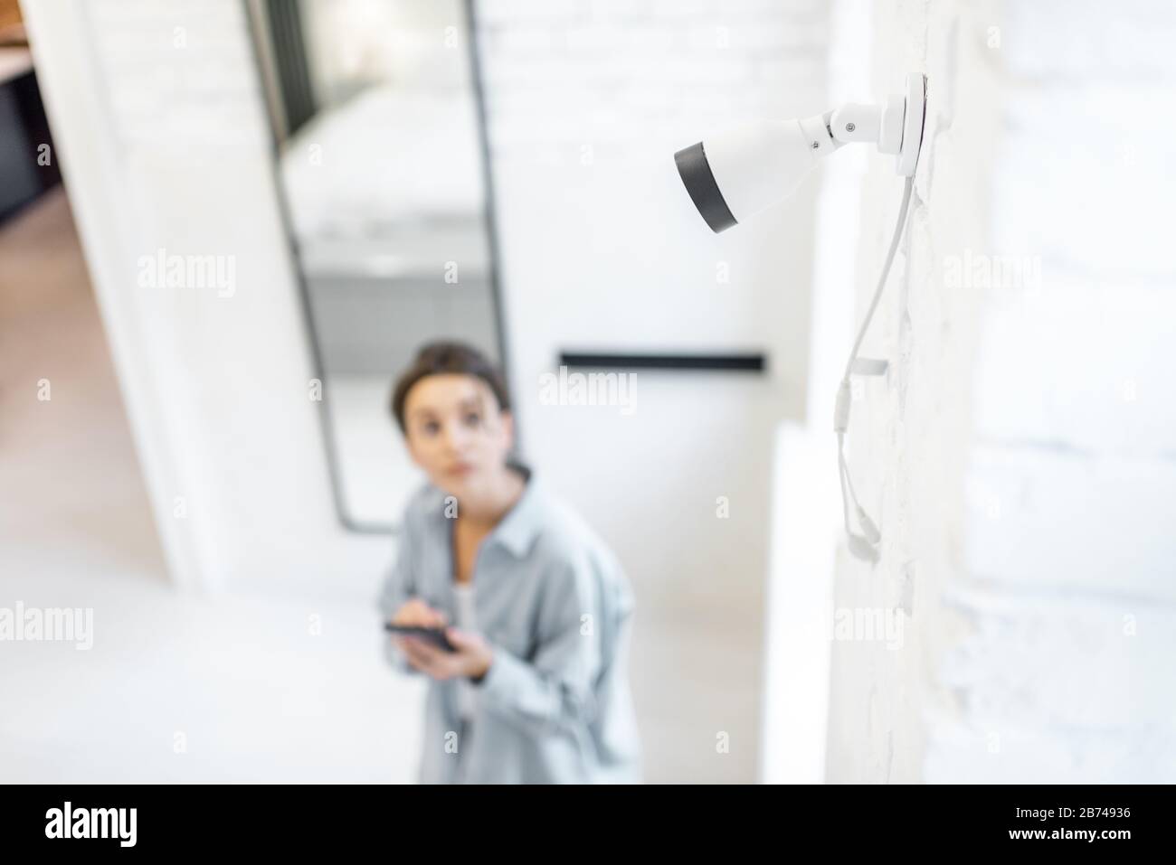 Woman controlla la videosorveglianza domestica con uno smartphone all'interno, la vista dall'alto e la telecamera ip in primo piano. Concetto di sistemi di sicurezza domestica wireless Foto Stock