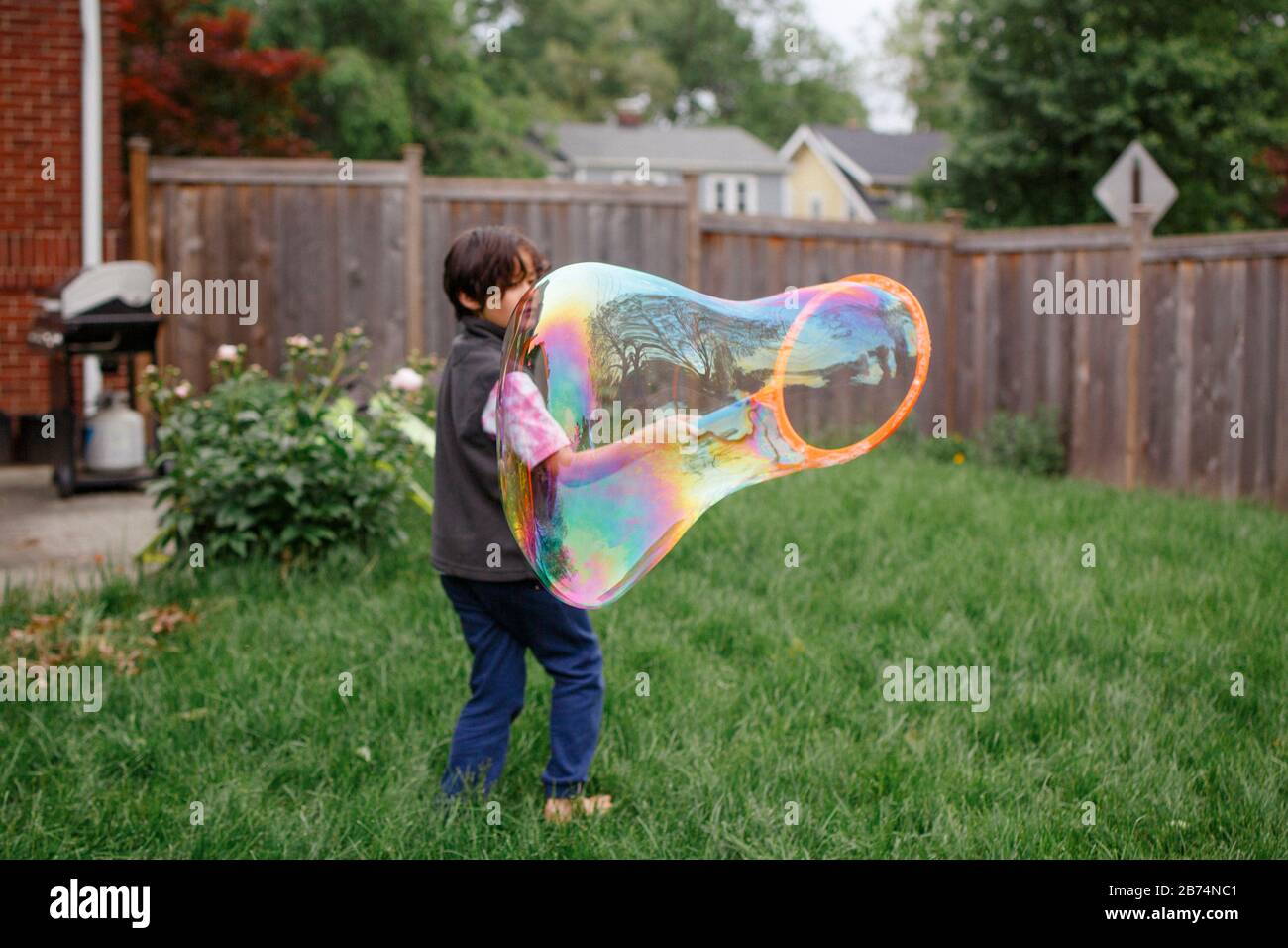 Un ragazzo a piedi nudi si alza a piedi nudi nel suo cortile creando una bolla gigante di sapone Foto Stock