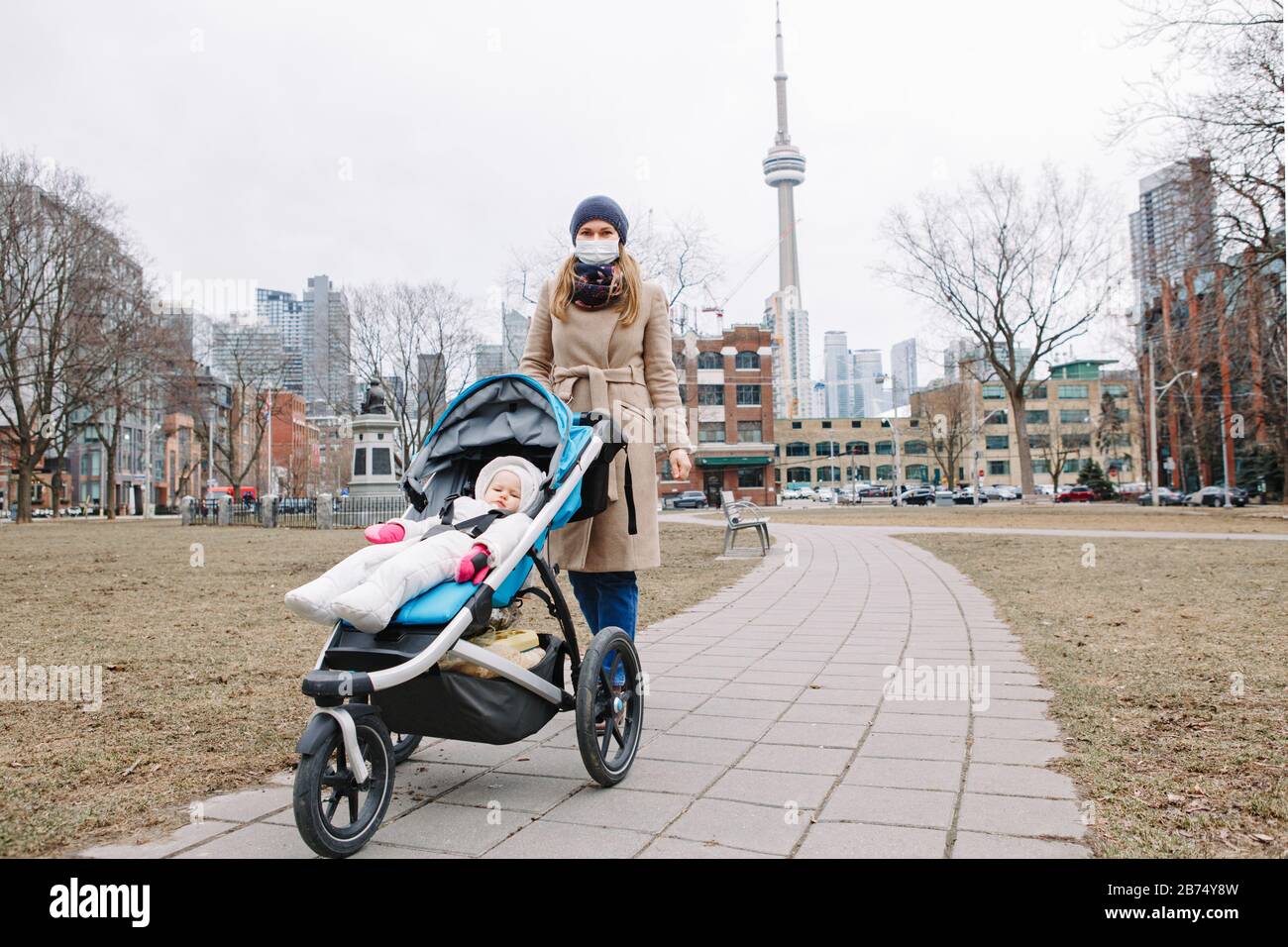 Giovane madre caucasica in maschera chirurgica camminare con il bambino all'aperto a Toronto. Maschera protettiva per il viso precauzione contro la nuova polmonite atipica cinese C Foto Stock