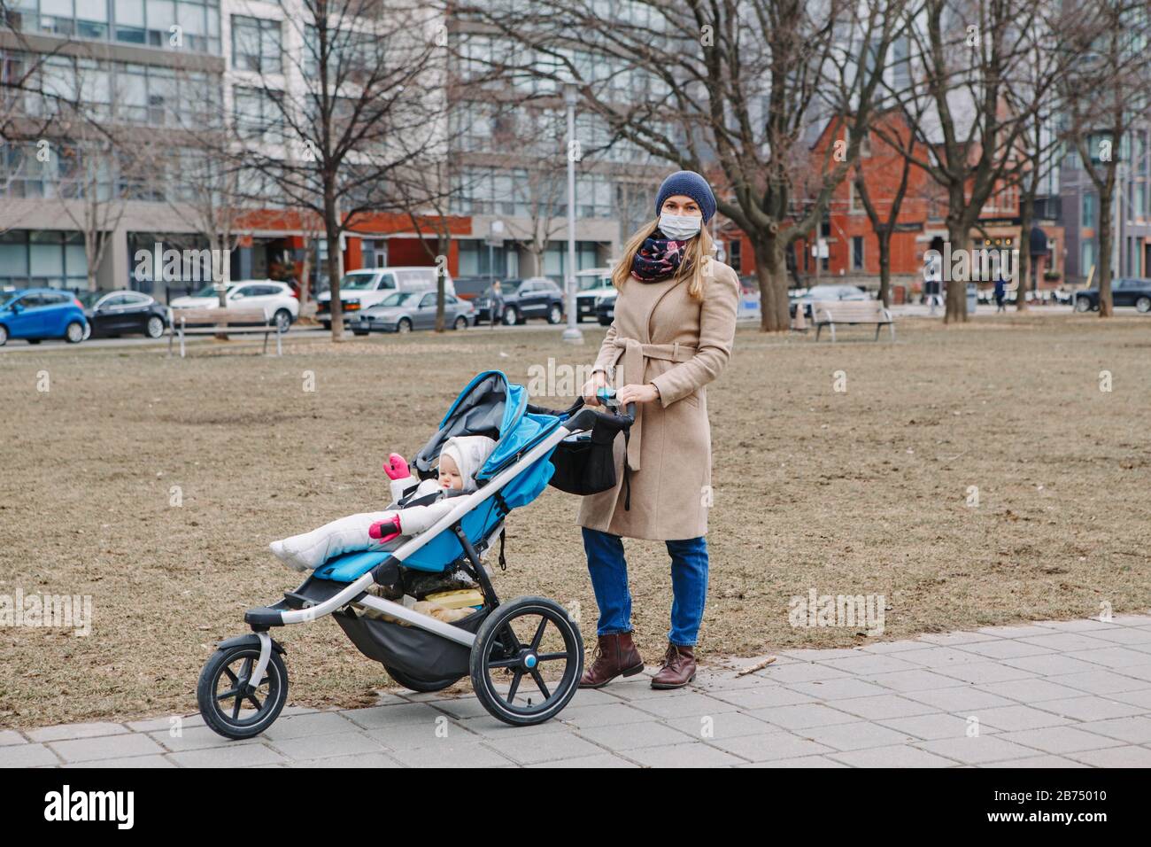 Giovane madre caucasica in maschera chirurgica camminare con il bambino all'aperto a Toronto. Maschera protettiva per il viso precauzione contro la nuova polmonite atipica cinese C Foto Stock