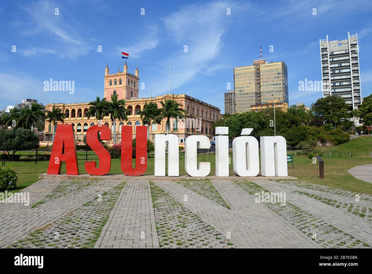 Cartello Asuncion di fronte al Palazzo del Governo in stile coloniale. Parco con alberi ed edifici del centro visibili. Popolare luogo di foto in Paraguay. Foto Stock