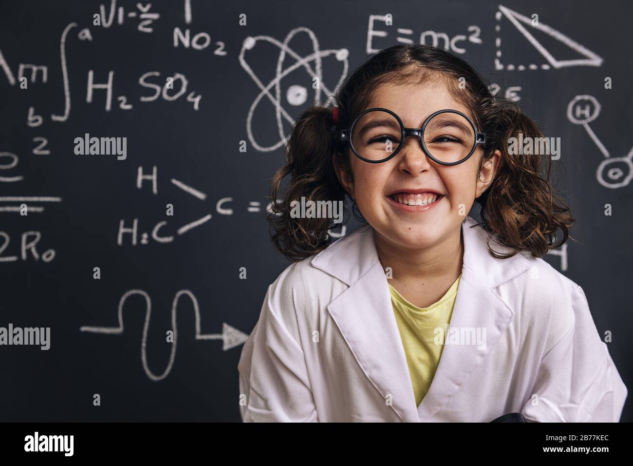 divertente bambina studente di scienza con occhiali in camice da laboratorio su sfondo lavagna scuola con disegni a mano scienza formula modello, ritorno a scuola Foto Stock