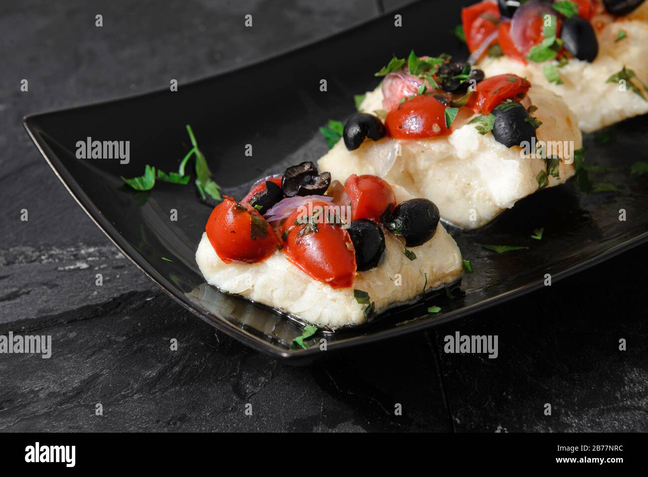 Filetti di merluzzo con olive nere, pomodori e cipolle su un piatto di ceramica nera - shot orizzontale da studio Foto Stock