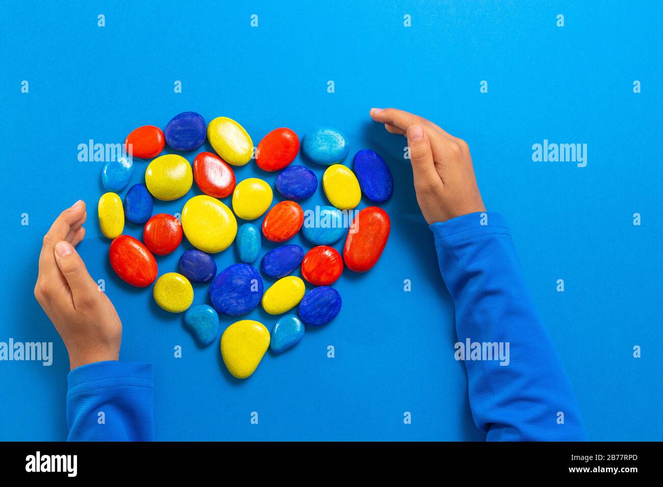 Concetto del giorno di consapevolezza di autismo del mondo. Mani del bambino con cuore fatte di pietre di colore rosso giallo blu su sfondo blu chiaro Foto Stock