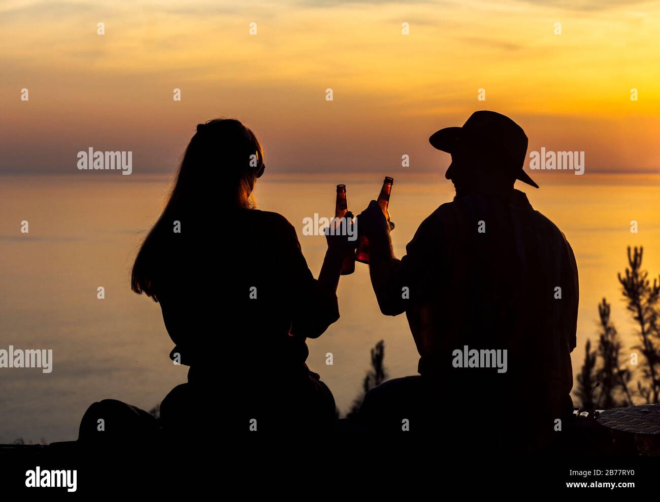 Silhouette di coppia romantica bere birra e sedersi su un bordo scogliera vicino al mare al tramonto. Foto Stock