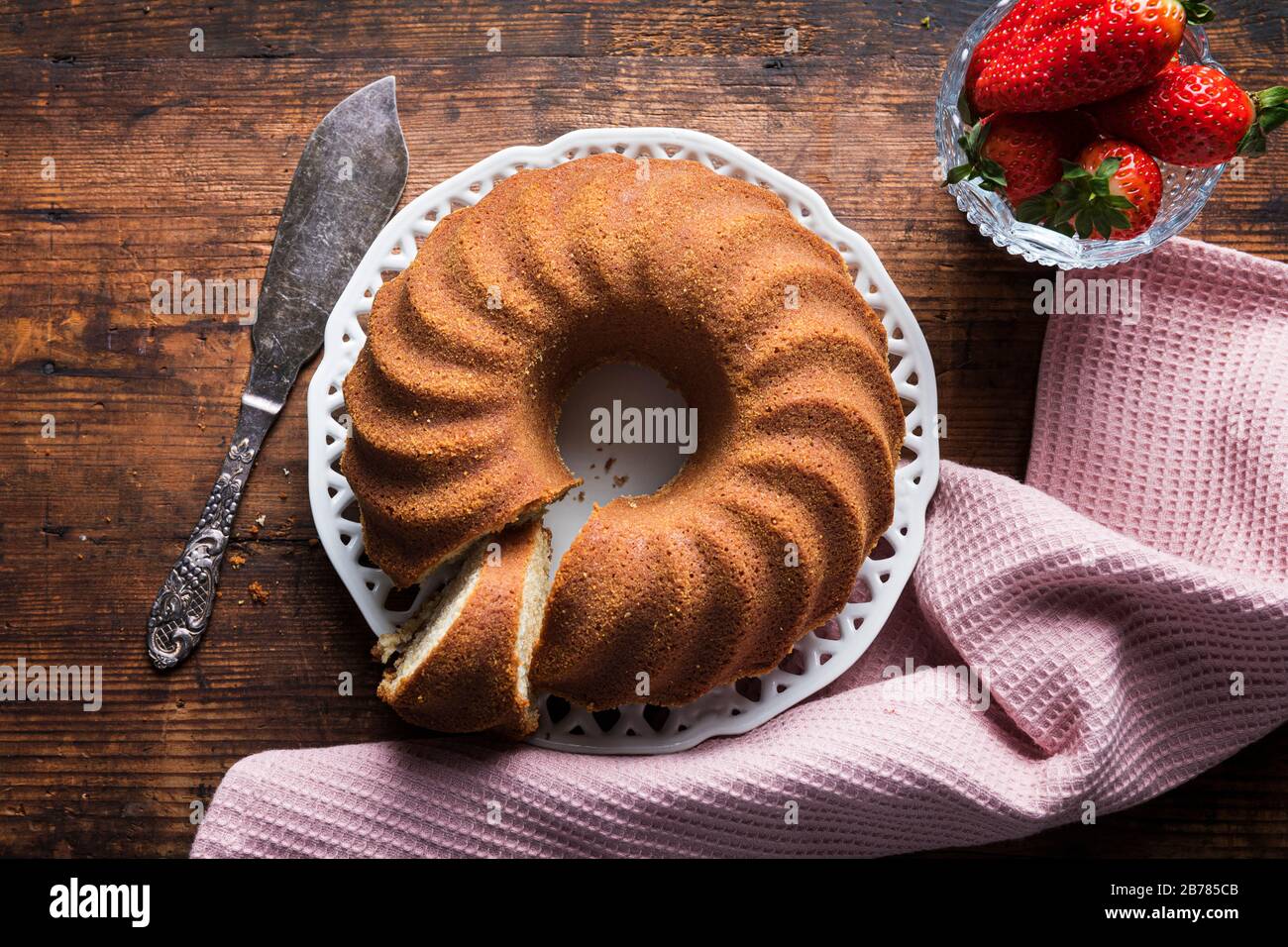 Kugelhupf, torta di impacchettamento o sockerkaka su una vecchia tabella rustica di legno. Con una ciotola di fragole, un tovagliolo rosa e una taglierina per torte d'annata. Visto da abov Foto Stock