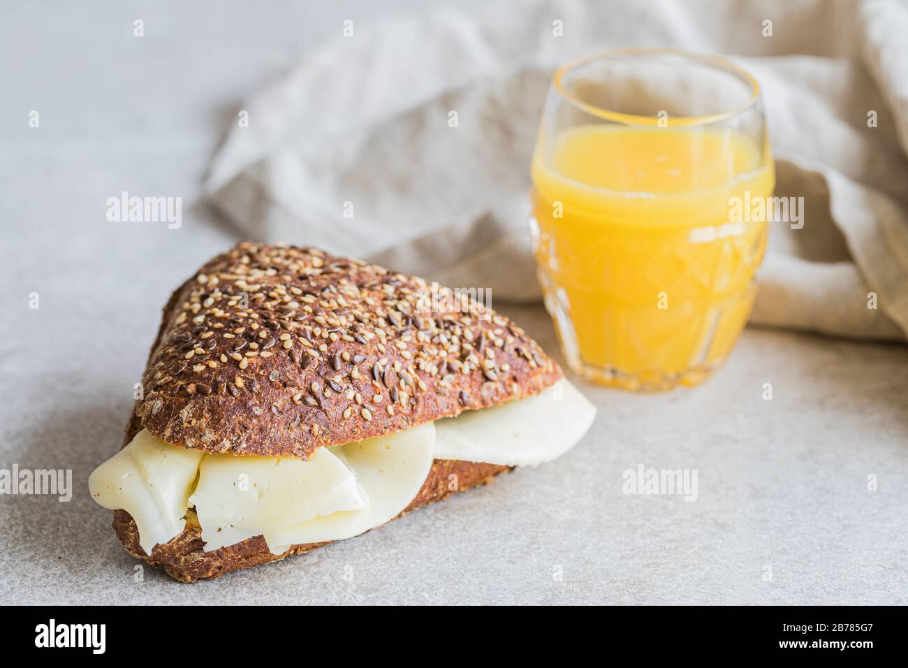 Un panino di formaggio sano fatto di pane organico scuro con i semi sulla parte superiore. Il pane è su un piccolo tavolo grigio con un bicchiere di succo d'arancia e una biancheria Foto Stock