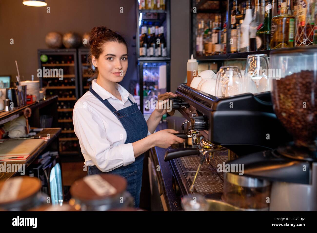 Una giovane cameriera di cafe vi guarda mentre siete in piedi accanto alla macchina del caffè e mentre preparano le bevande per i clienti al lavoro Foto Stock
