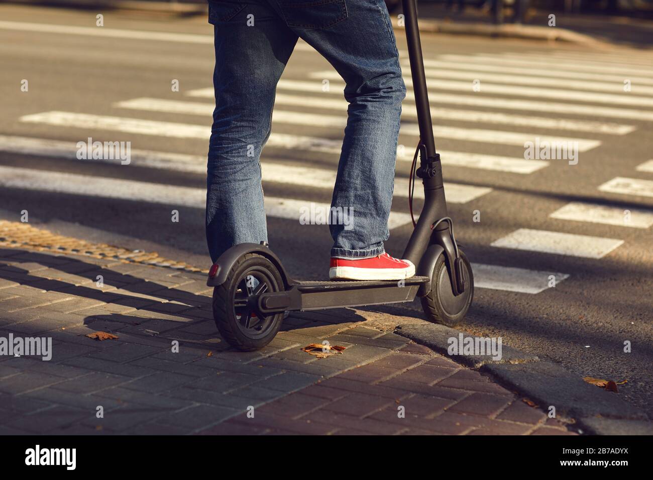 Gambe di un uomo in jeans e scarpe da ginnastica su uno scooter elettrico a una passeggiata a piedi su una strada della città Foto Stock