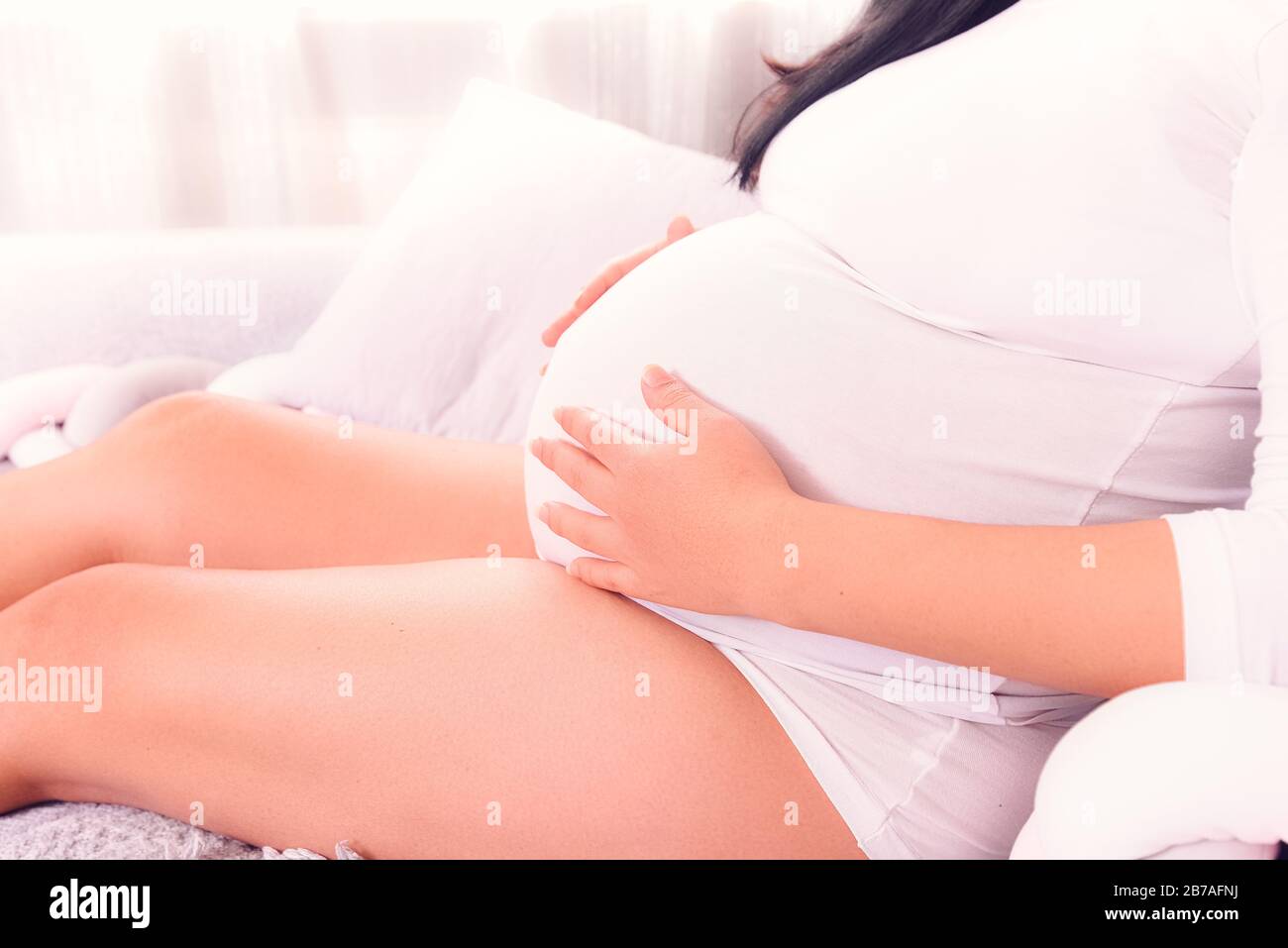 Donna incinta seduta sul divano e tenendo il bambino incinta con due mani con la parte superiore. Tempo di maternità. Foto Stock