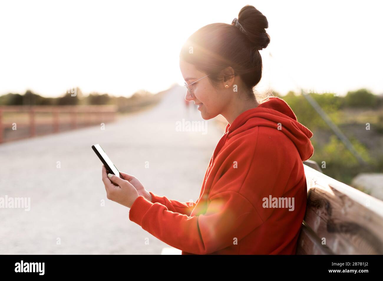 Giovane donna con gli occhiali sui suoi venti anni che chiacchierano sul suo telefono mentre siede in una panchina di un parco. Sorride e indossa un maglione rosso e un pony Foto Stock