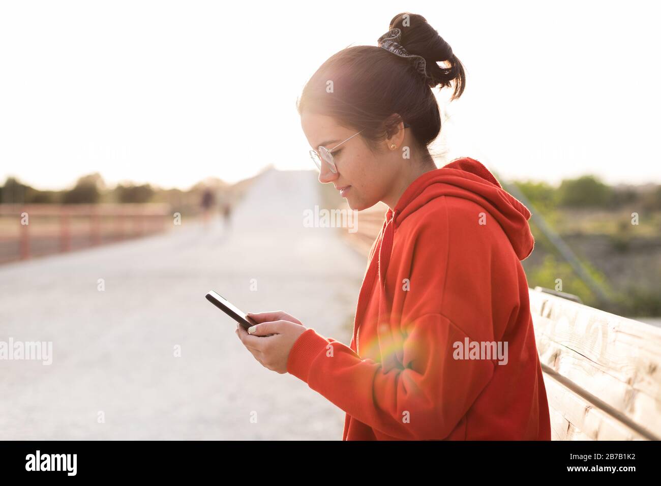 Giovane donna con gli occhiali sui suoi venti anni che chiacchierano sul suo telefono mentre siede in una panchina di un parco. Sorride e indossa un maglione rosso e un pony Foto Stock