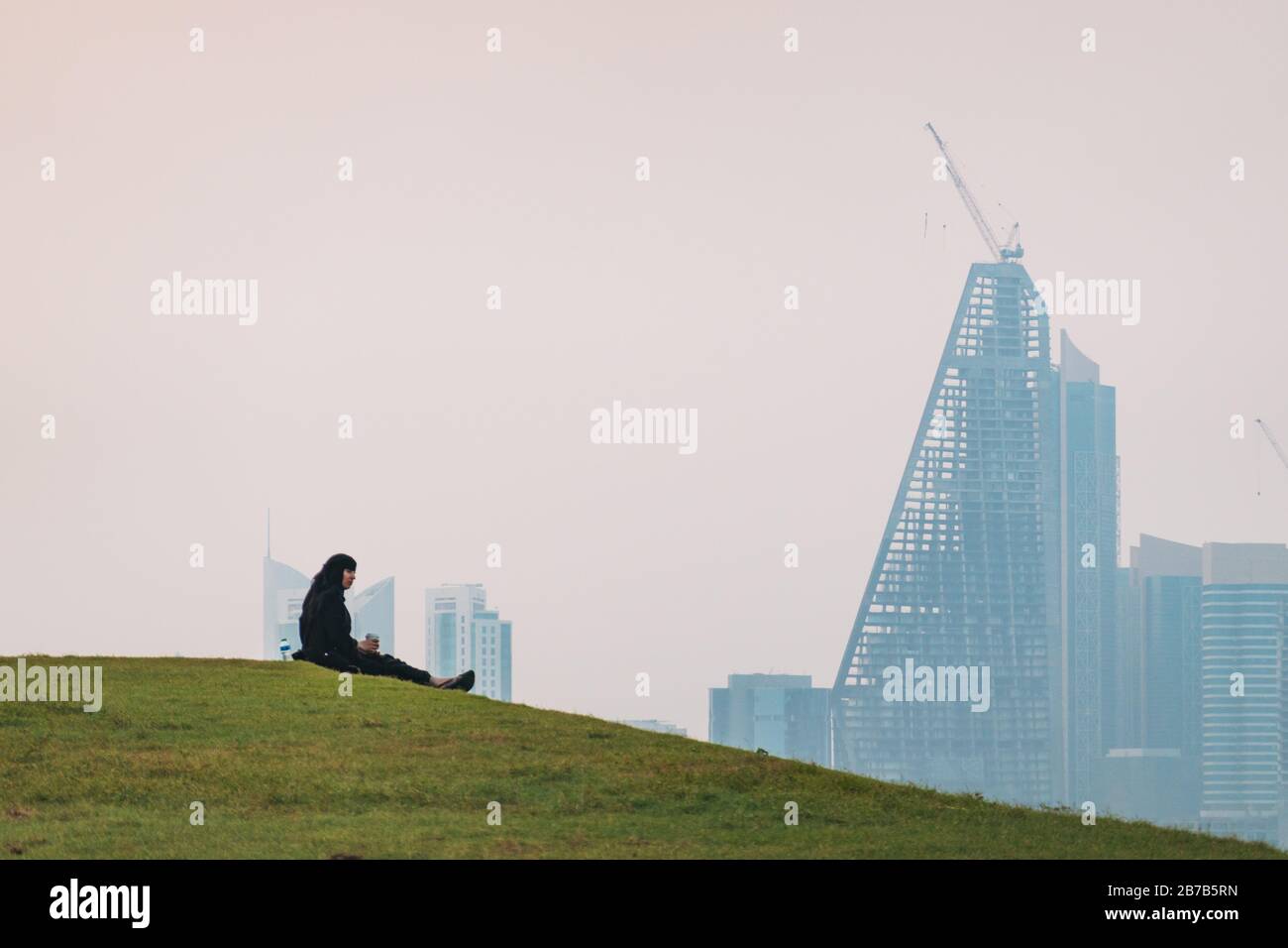 Una donna musulmana ama rinfreschi seduti su una collina erbosa a Doha, Qatar. Lo skyline del quartiere finanziario può essere visto attraverso la foschia sullo sfondo Foto Stock
