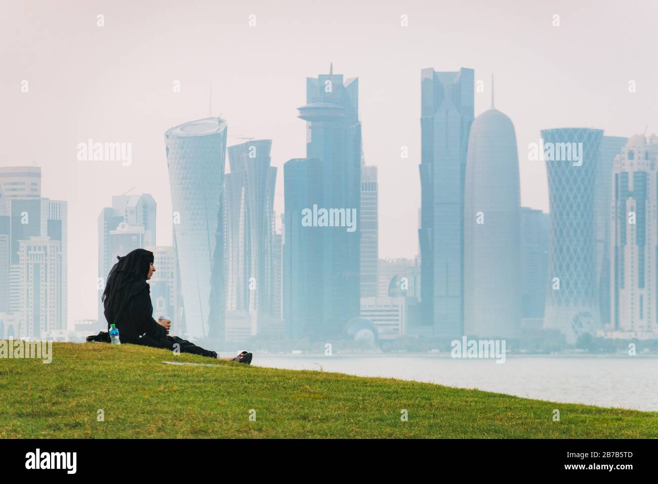 Una donna musulmana ama rinfreschi seduti su una collina erbosa a Doha, Qatar. Lo skyline del quartiere finanziario può essere visto attraverso la foschia sullo sfondo Foto Stock