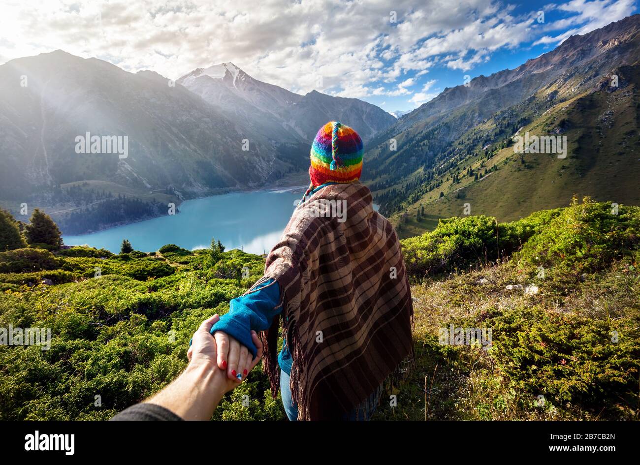 Donna turistica in cappello arcobaleno e poncho marrone tenendo l'uomo a mano e andando al lago in montagna Foto Stock